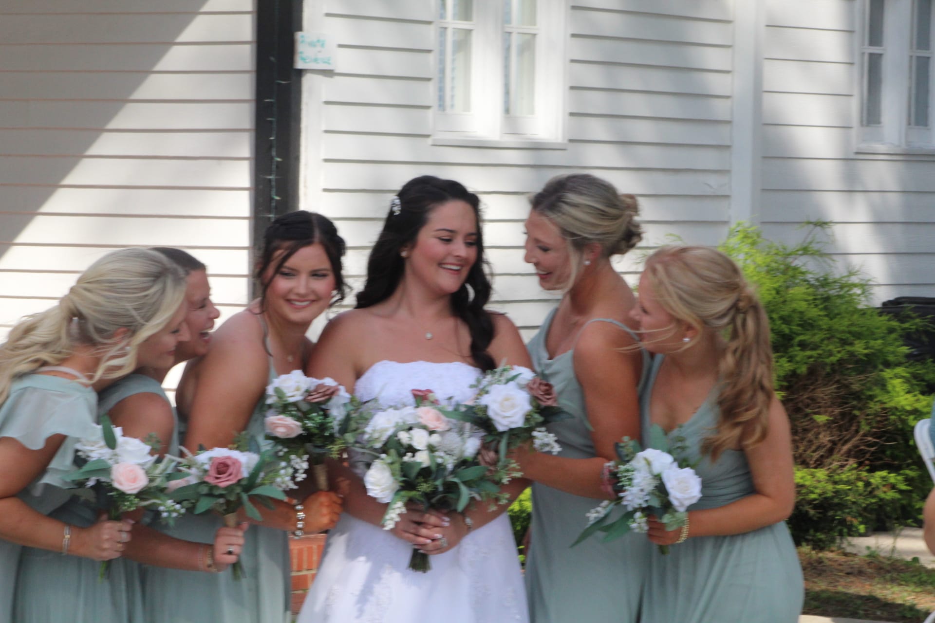 Bride in a strapless white gown laughs with four bridesmaids in sage green dresses holding white and blush floral bouquets outside a historic white clapboard building.