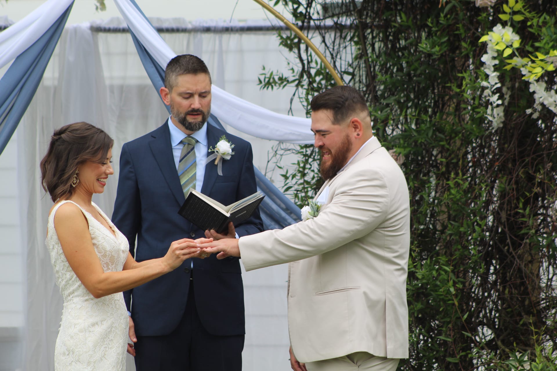 Bride smiling as groom places wedding ring on her finger during outdoor ceremony beneath a draped floral arch at Scarborough House.