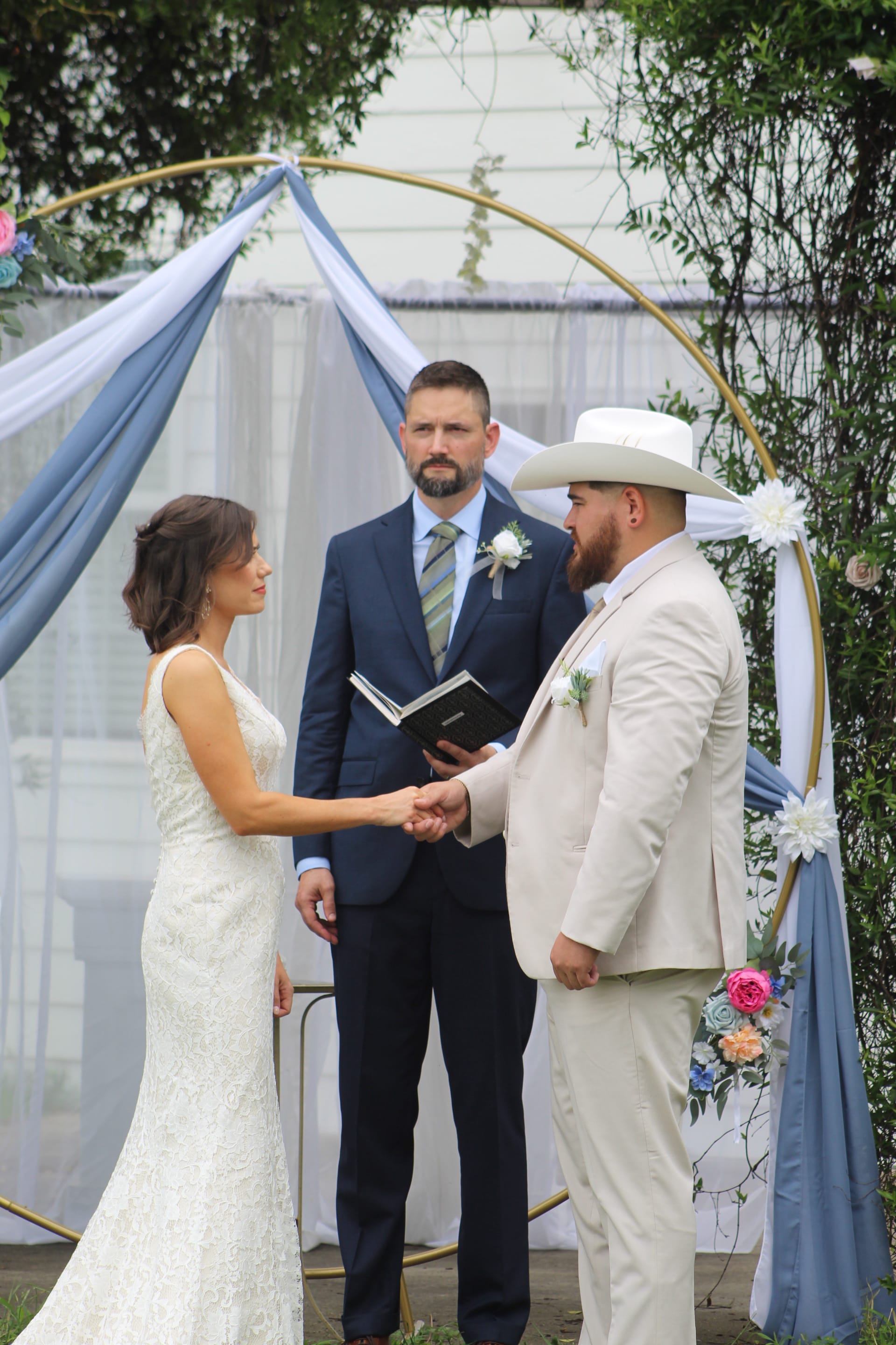 Bride in lace gown and groom in cream suit and white cowboy hat hold hands during outdoor wedding ceremony beneath a gold circular arch draped in dusty blue fabric at Scarborough House.