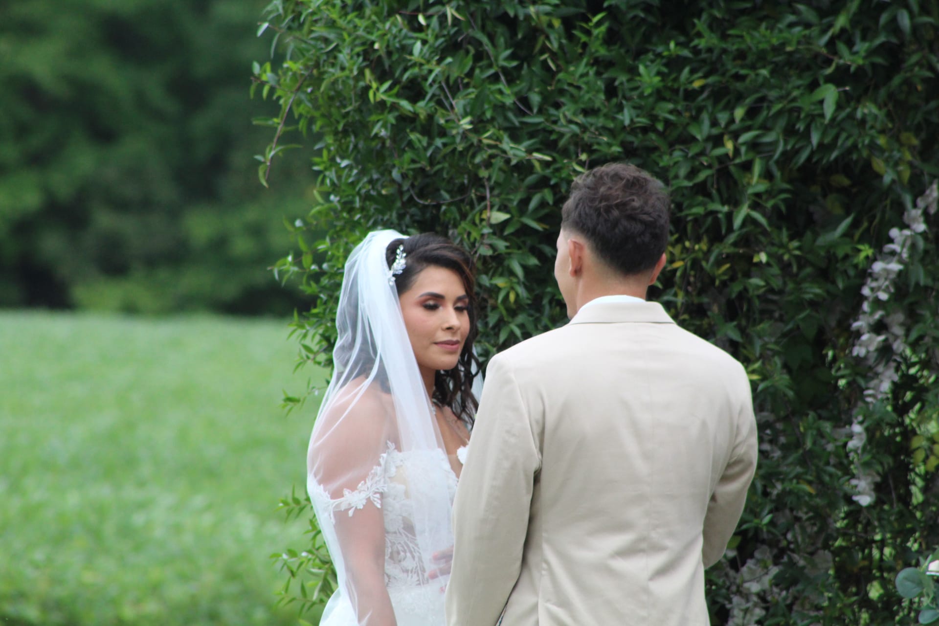 Bride in lace gown and white veil gazes softly at her groom during an outdoor garden ceremony at Scarborough House estate surrounded by lush green foliage.