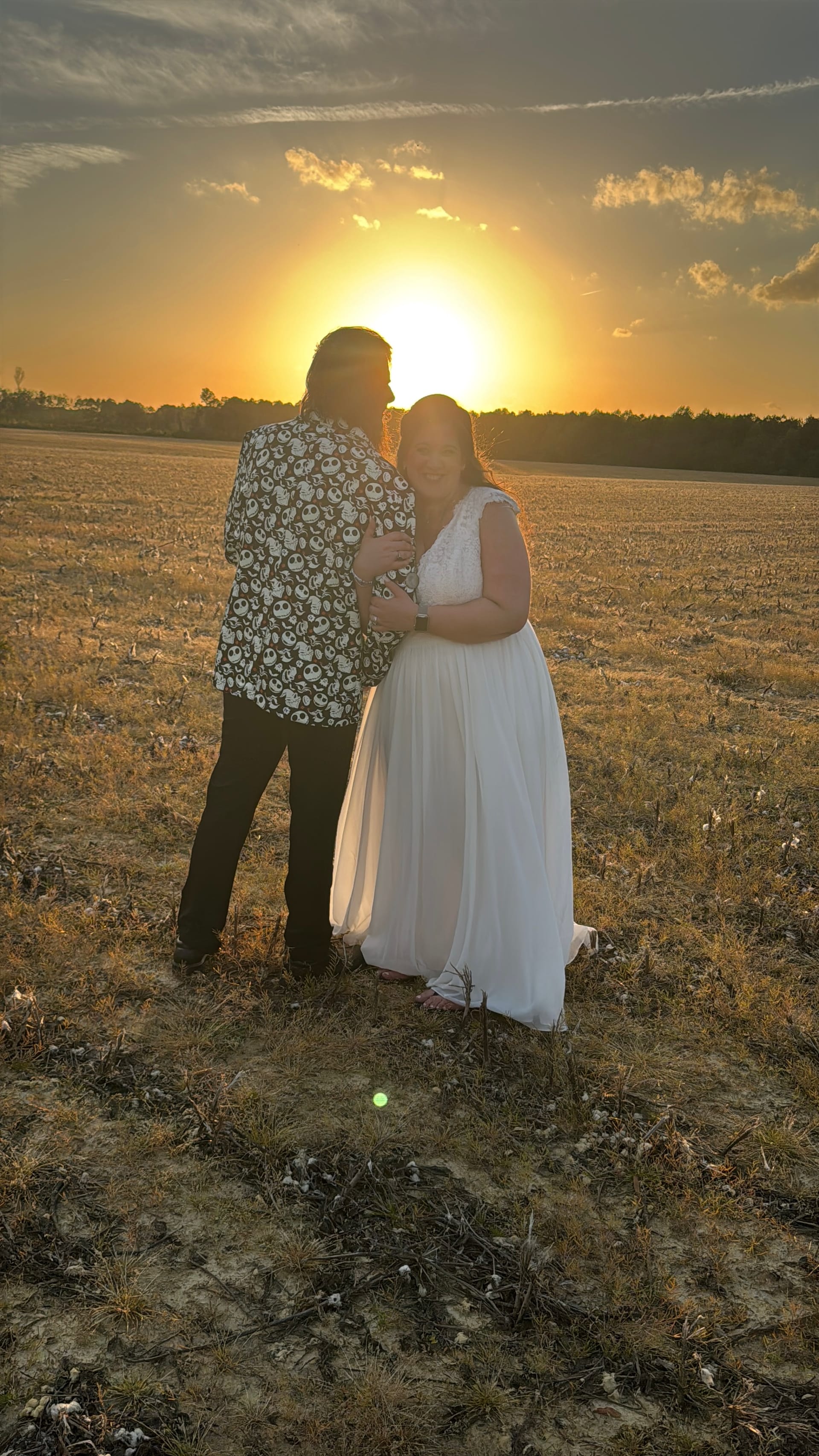 Bride in a flowing white gown and groom in a patterned shirt embrace in an open farmland field at golden hour with a glowing sunset behind them at Scarborough House estate.