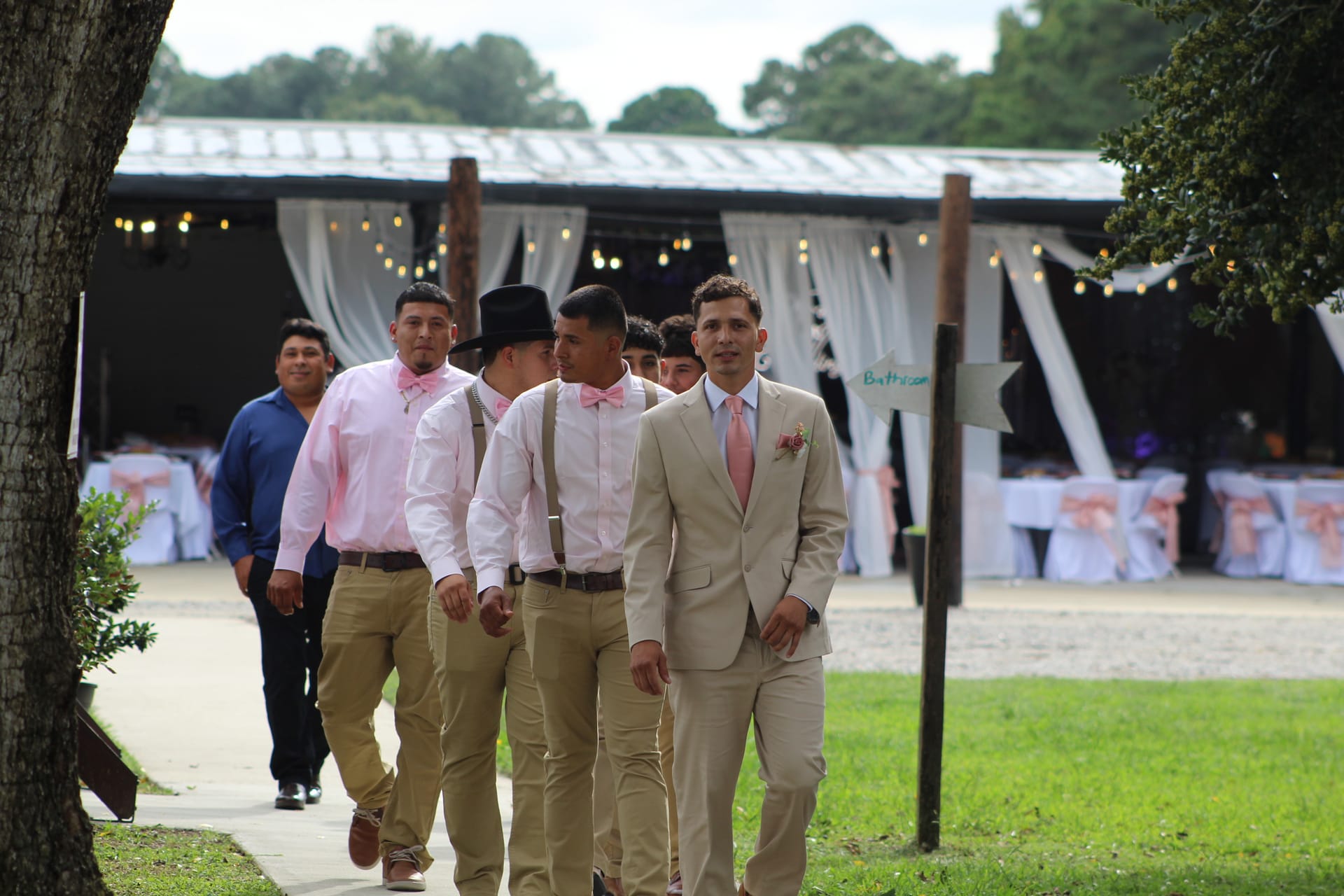 Groom in a tan suit with pink tie leads his groomsmen in pink shirts and khaki pants along a path toward the decorated outdoor pavilion at Scarborough House wedding venue.