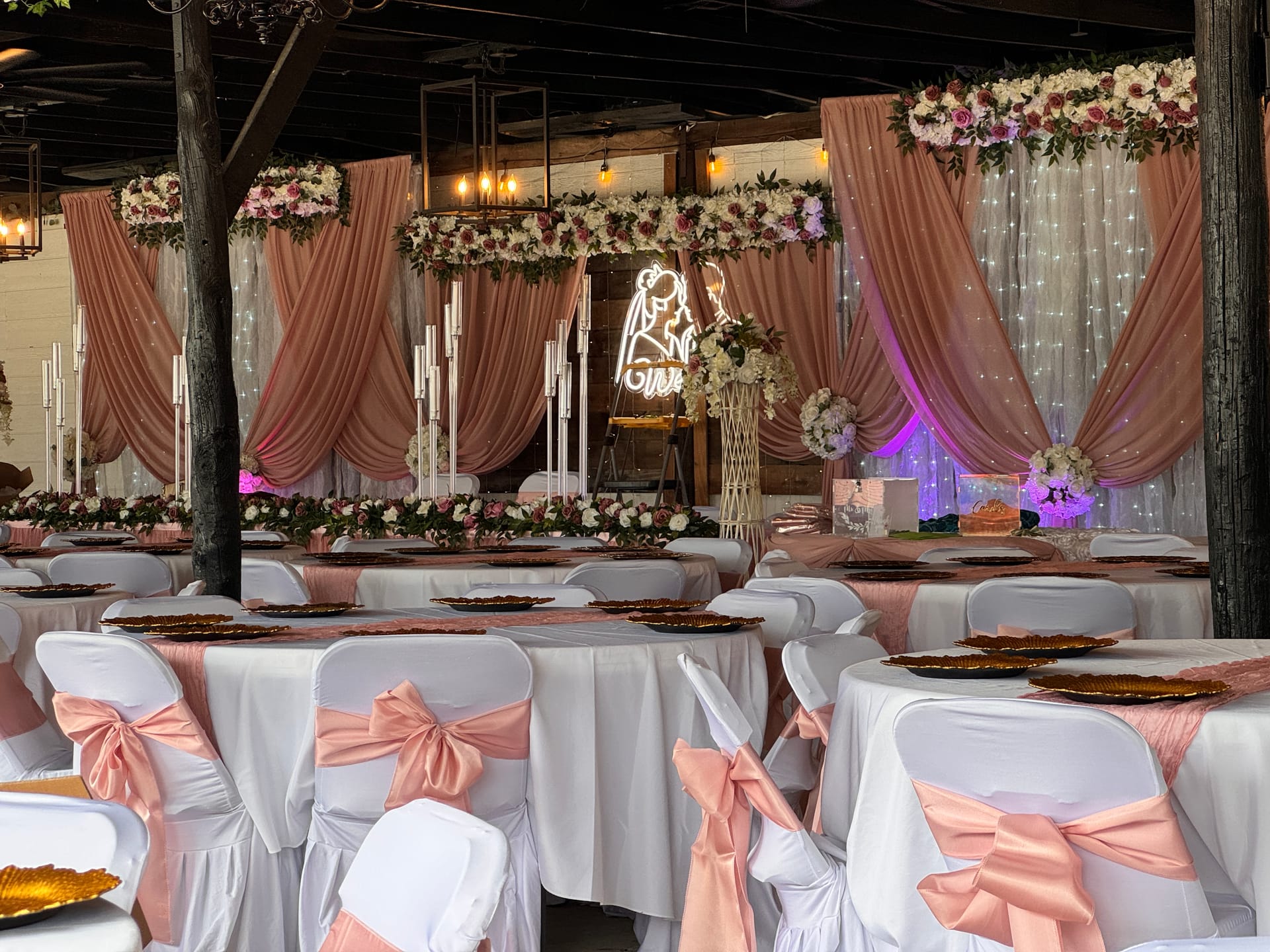 Elegantly decorated wedding reception hall featuring blush pink draped fabric backdrop with floral garlands, white chair covers with pink sashes, gold charger plates, and a glowing neon bride and groom sign at Scarborough House.