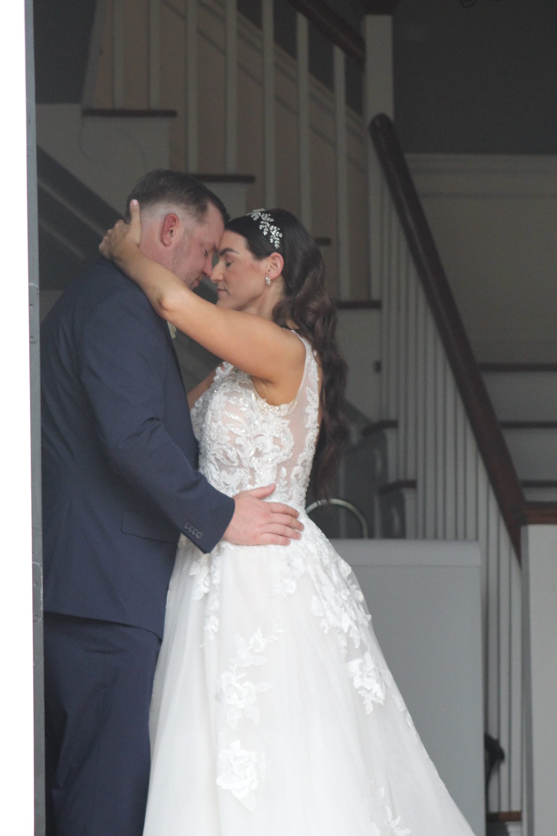 Bride in a lace ballgown and groom in a navy suit share an intimate forehead-to-forehead embrace inside the grand hall staircase of Scarborough House historic estate.