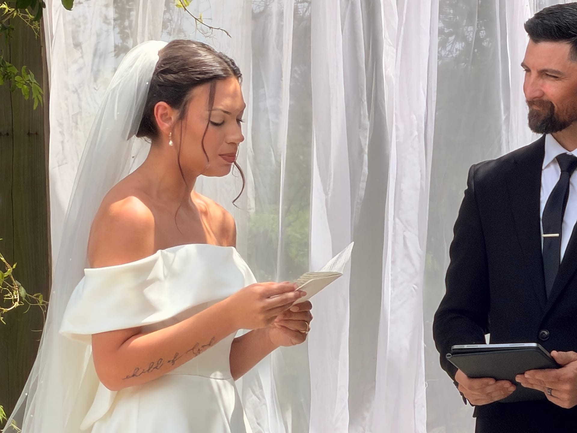 Bride in an off-shoulder white gown and veil reads her handwritten vows aloud as the suited groom watches with a warm smile during an outdoor wedding ceremony.