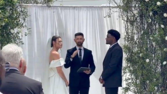 Bride in an off-shoulder white gown and groom in a dark suit exchange vows before an officiant beneath a white draped backdrop adorned with greenery and white florals at Scarborough House.