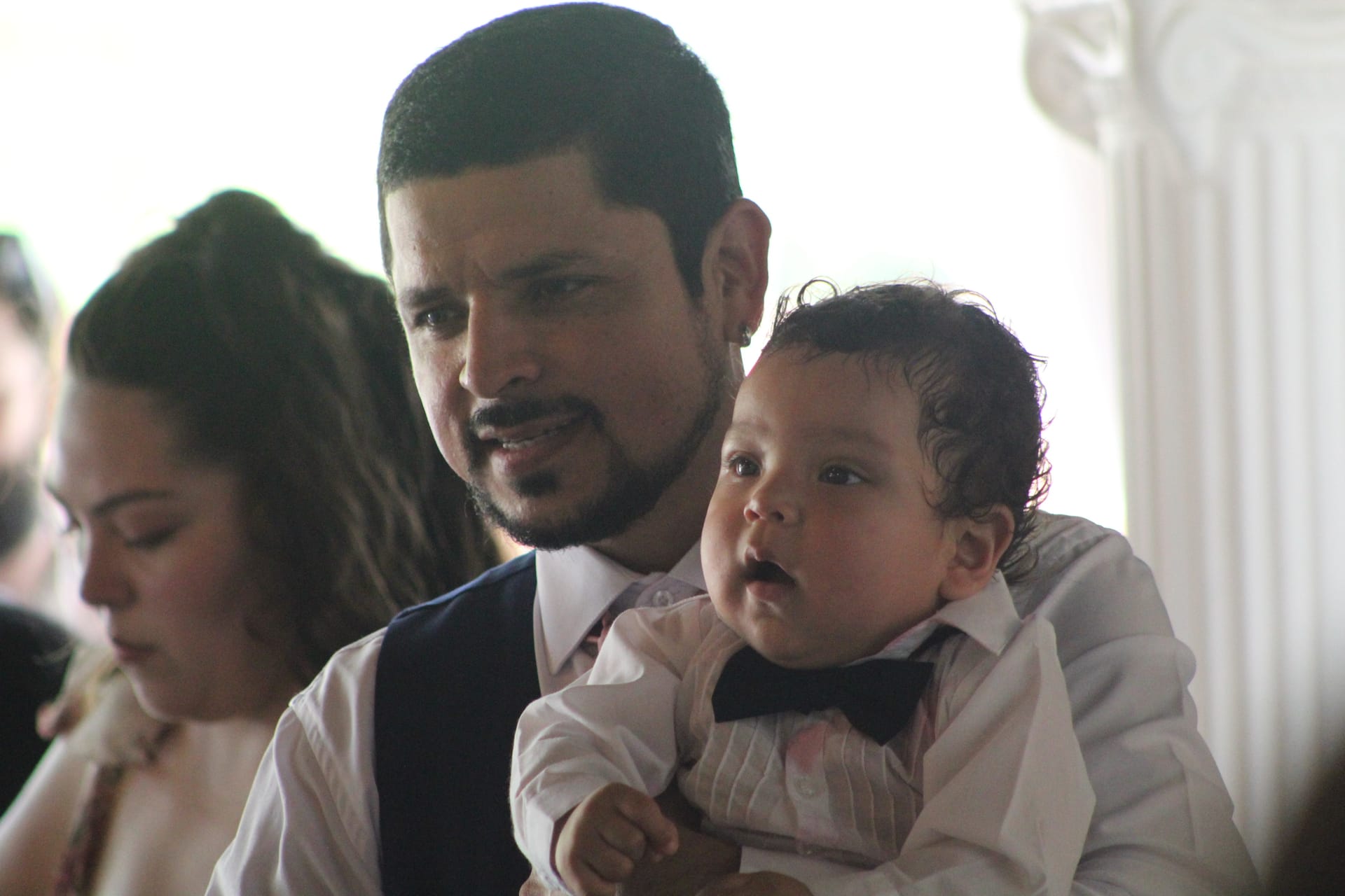 A well-dressed father in a vest and white shirt holds his infant dressed in a matching white shirt and bow tie during a wedding reception at Scarborough House.