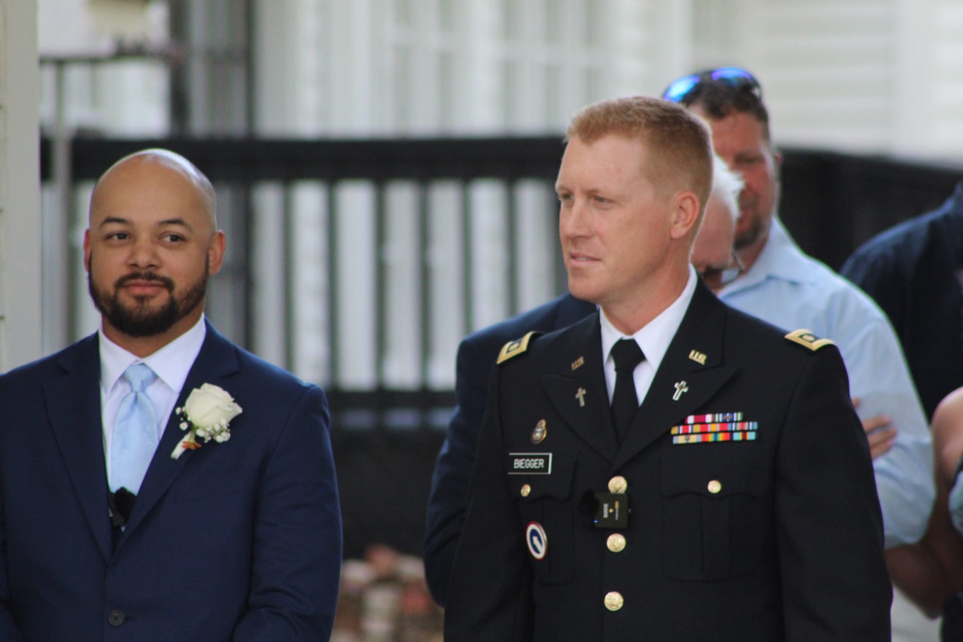 A US Army soldier in dress uniform stands beside a groomsman in a navy suit with a white boutonniere at an outdoor wedding venue.