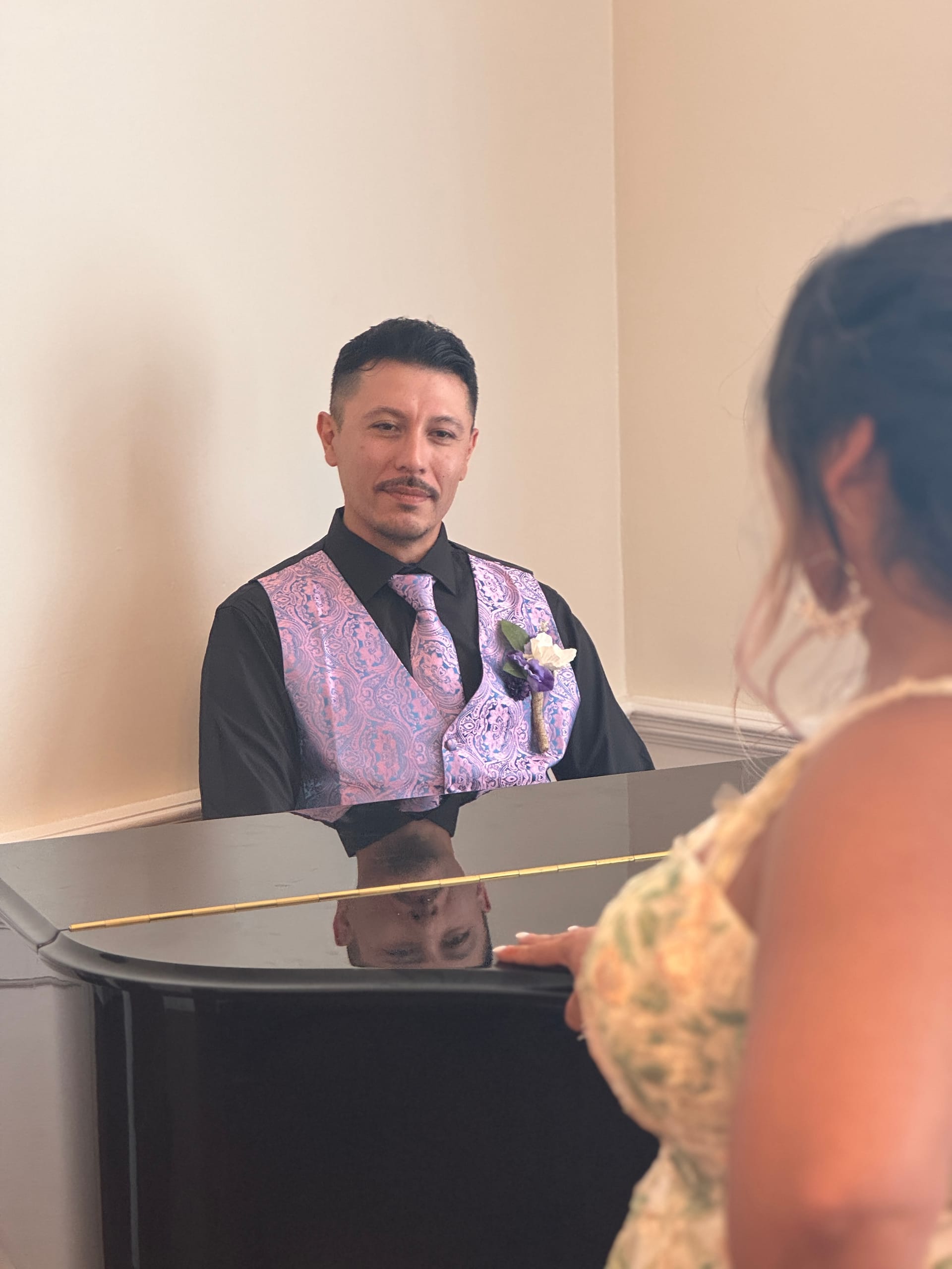 Groom in a black shirt and lavender paisley vest with boutonniere stands behind a grand piano in the historic grand hall, his reflection visible in the polished lid as a guest looks on.