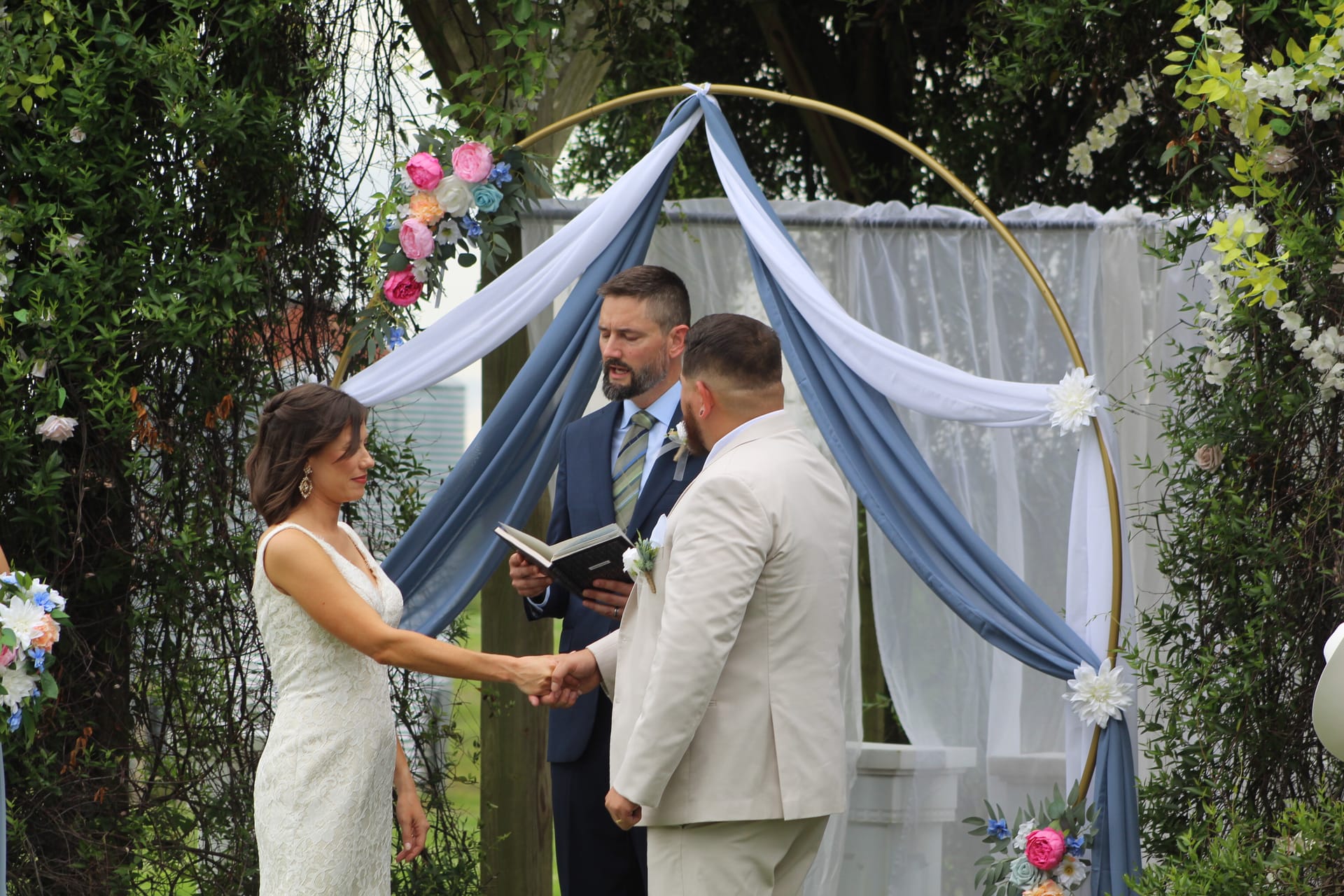 Bride and groom hold hands during outdoor wedding vows beneath a gold circular arch draped in dusty blue and white fabric adorned with colorful florals at Scarborough House.