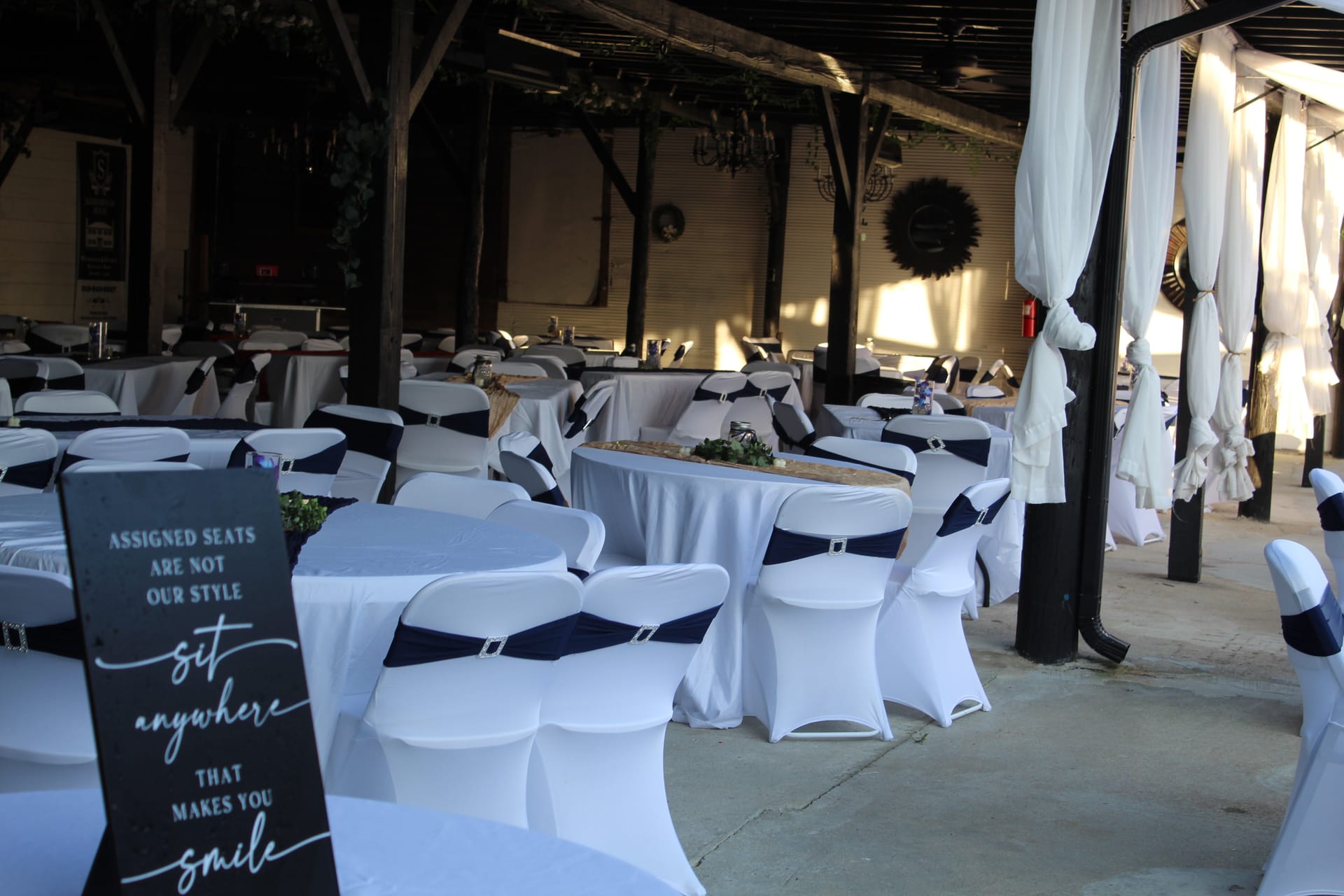 Magnolia pavilion reception setup at Scarborough House featuring white spandex chair covers with navy sashes, round tables with greenery centerpieces, draped white curtain columns, and a chalkboard open-seating sign in the foreground.