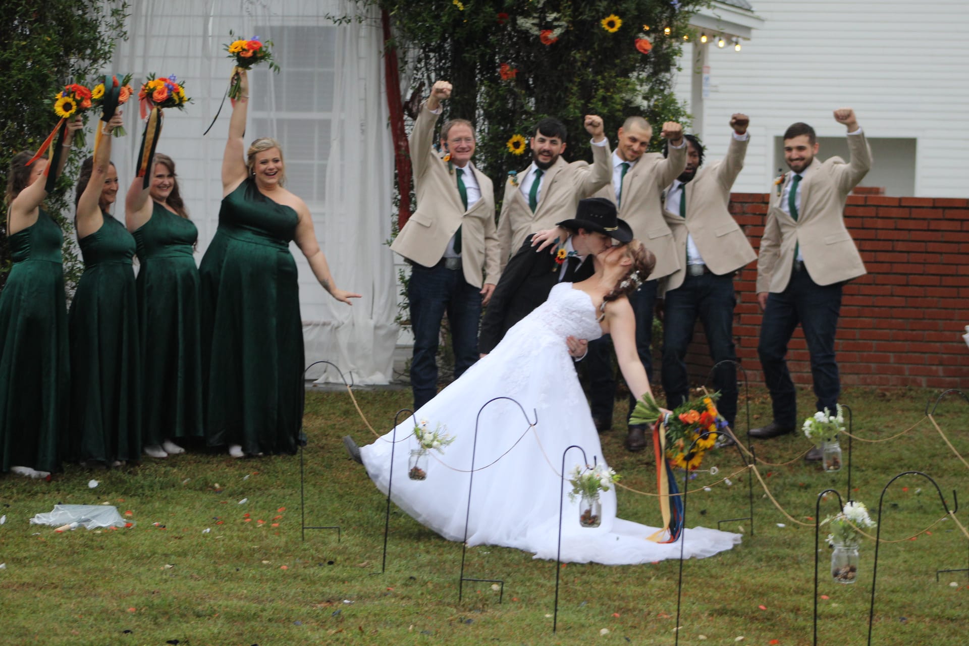 Groom dips bride for a kiss on the ceremony lawn while bridesmaids in emerald green gowns and groomsmen in tan blazers cheer behind them at Scarborough House.