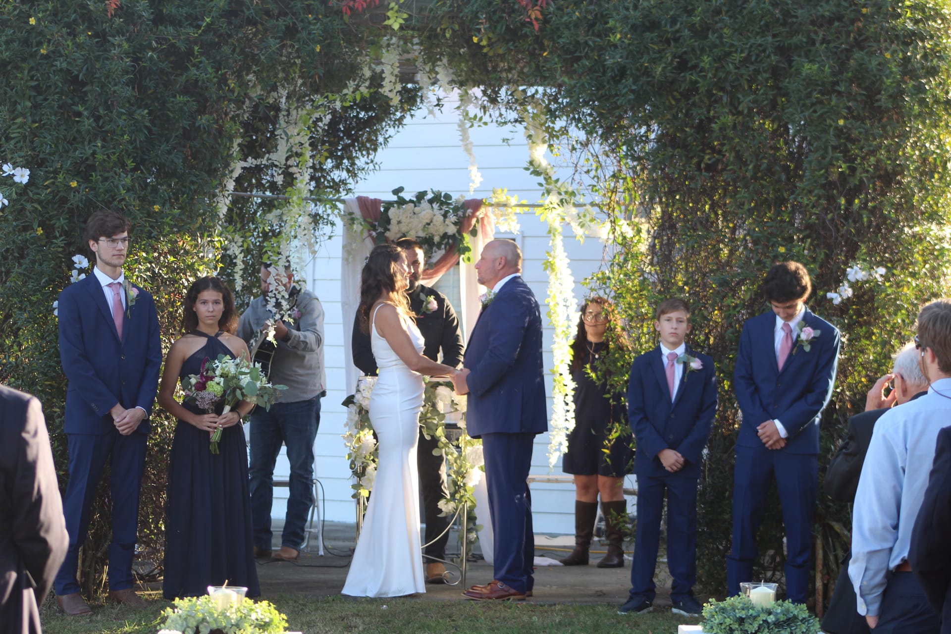 Bride and groom exchange vows beneath a lush green arbor adorned with white florals and cascading greenery at an outdoor wedding ceremony, surrounded by their wedding party.