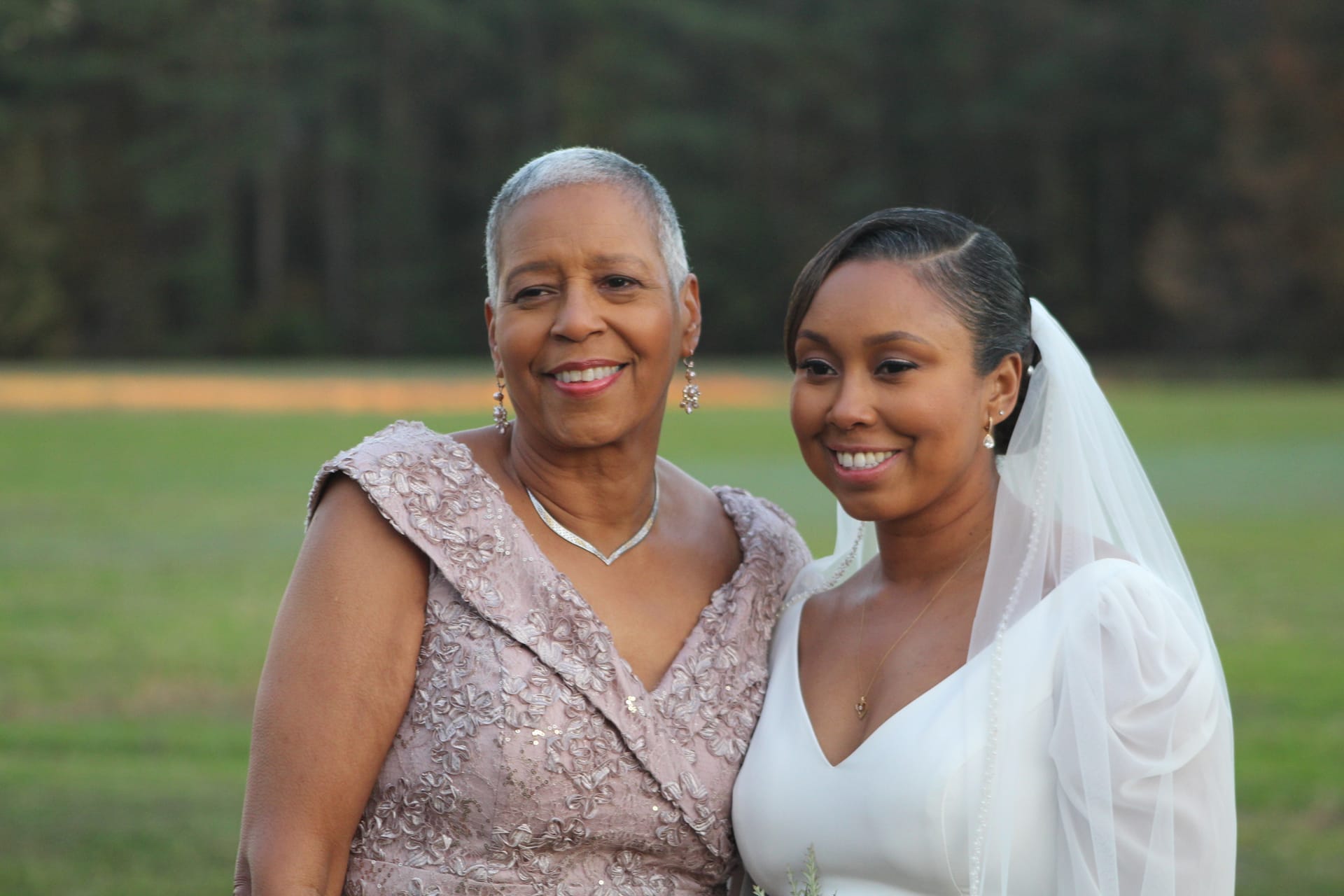 A bride in a white gown and veil smiles alongside an older woman in a mauve lace dress against a soft backdrop of green fields and trees at Scarborough House.