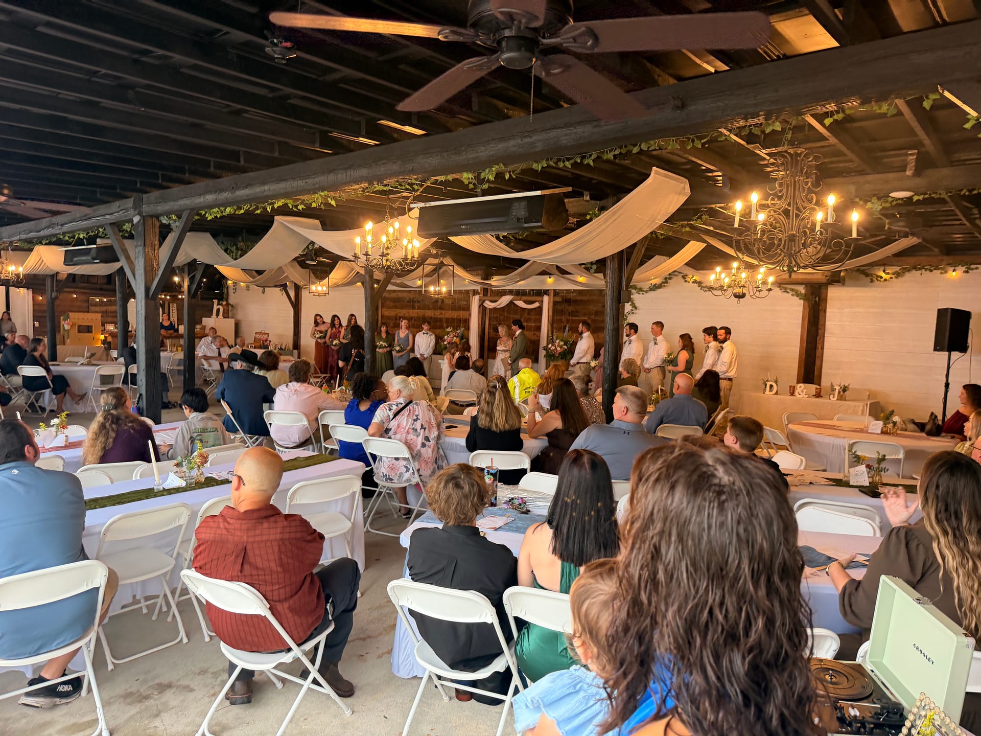 Guests seated at round tables watch a wedding ceremony under a rustic wood-beam pavilion adorned with draped fabric, chandelier lighting, and greenery at Scarborough House.