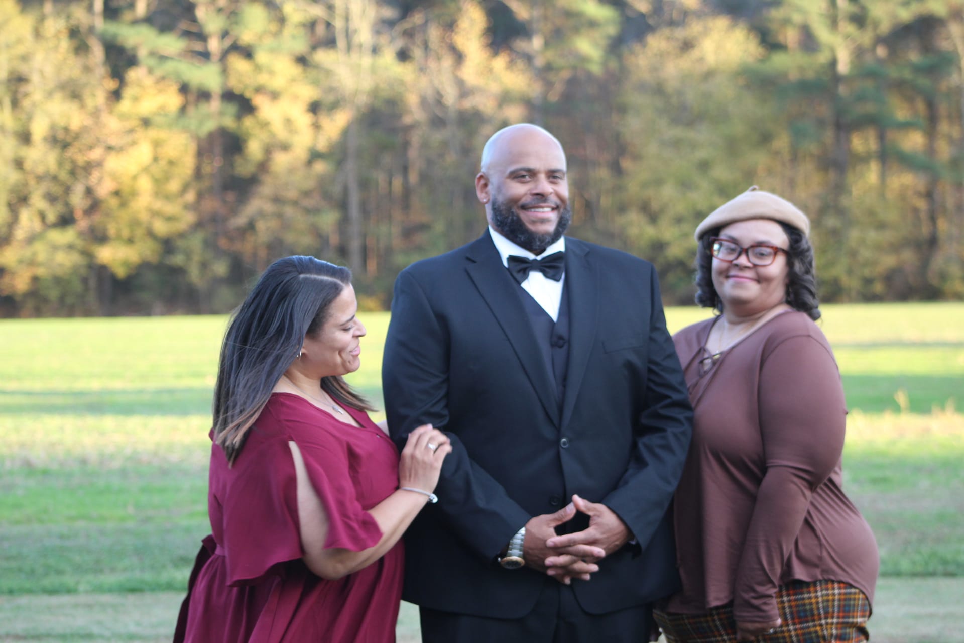 A smiling man in a black tuxedo stands between two women on a sunlit lawn at Scarborough House with golden autumn trees in the background.