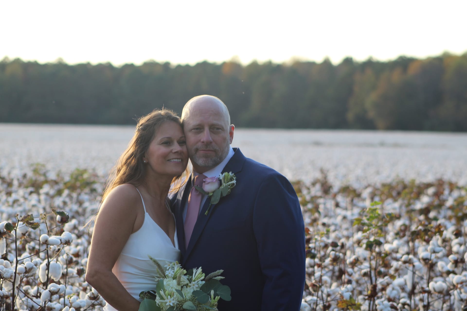 Bride in white gown and groom in navy suit with pink boutonniere pose together in a cotton field at golden hour with a tree line glowing in the background at Scarborough House.
