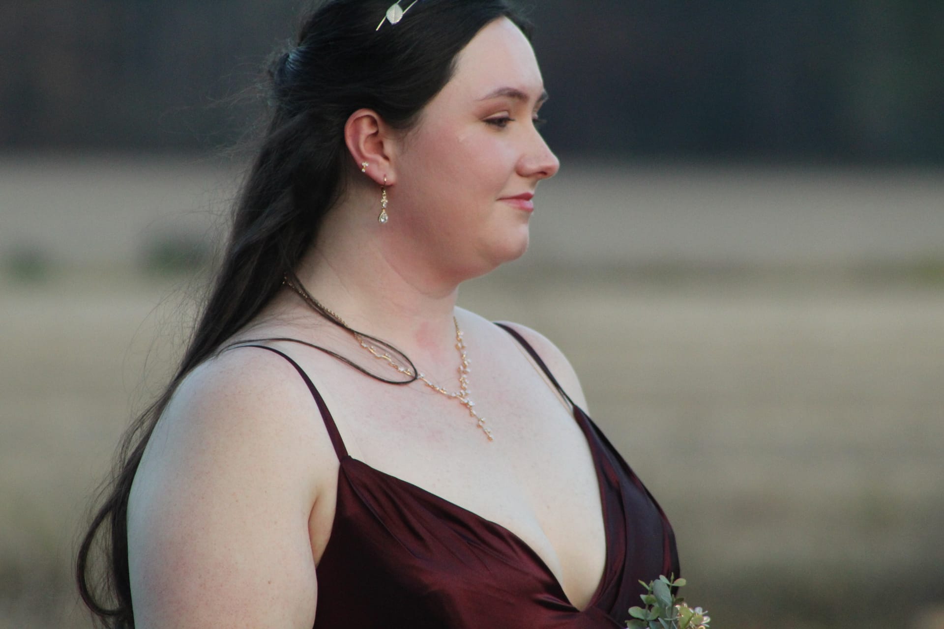 A bridesmaid in a deep burgundy gown with gold jewelry gazes softly downward against a blurred golden farmland backdrop at Scarborough House.