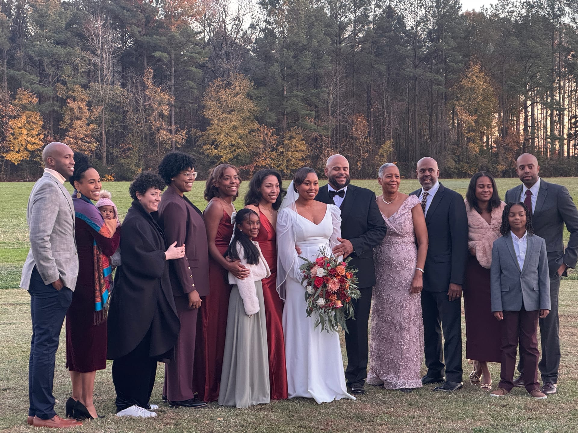 Bride in white gown holding a fall bouquet and groom in black tuxedo pose with their extended family on the open farmland fields at Scarborough House during golden hour surrounded by autumn trees.