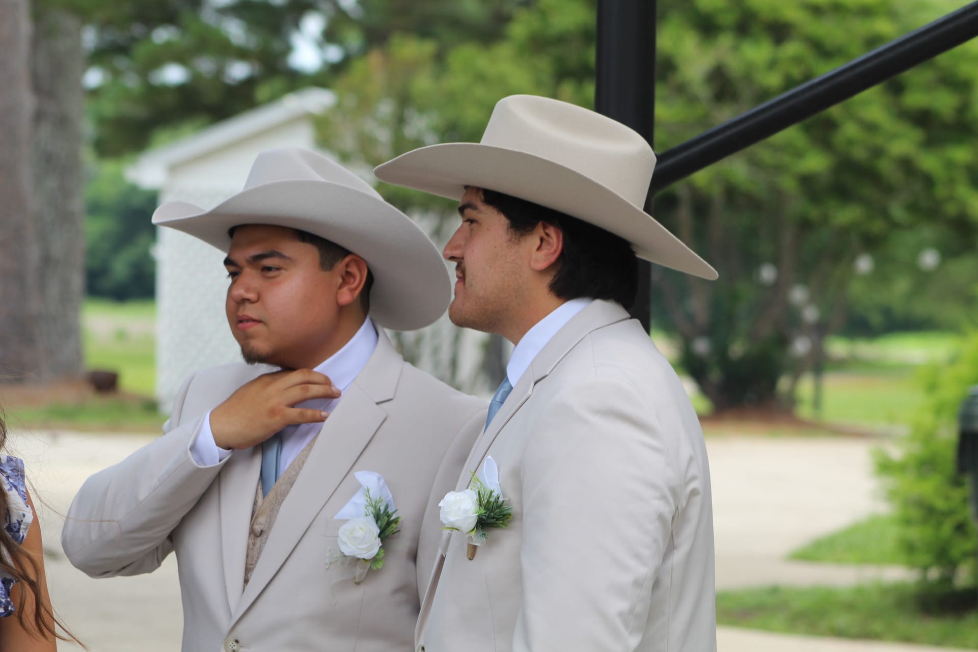 Two groomsmen in tan suits and cowboy hats adjust each other's ties outdoors before a rustic wedding ceremony at Scarborough House.