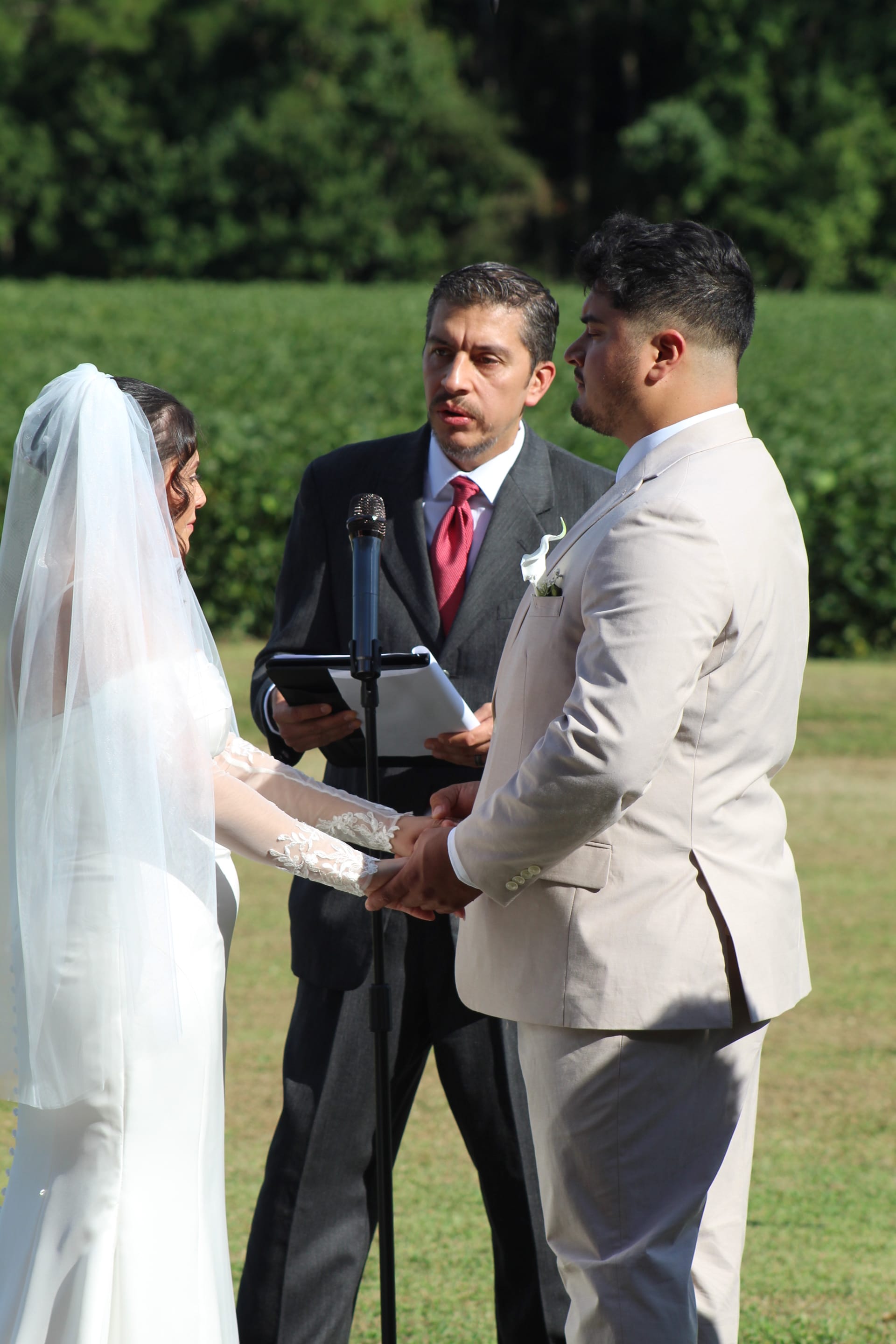 Bride and groom hold hands during outdoor wedding vows as officiant reads from a tablet on a sunlit ceremony lawn surrounded by lush green fields and trees.