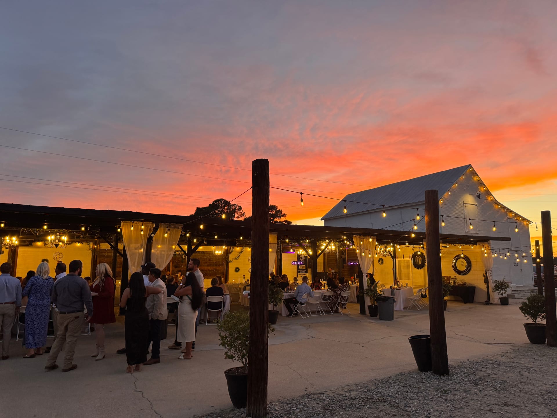 Guests mingle on a warmly lit outdoor patio at Scarborough House wedding venue beneath a breathtaking orange and pink sunset sky with string lights glowing across the white barn structure.