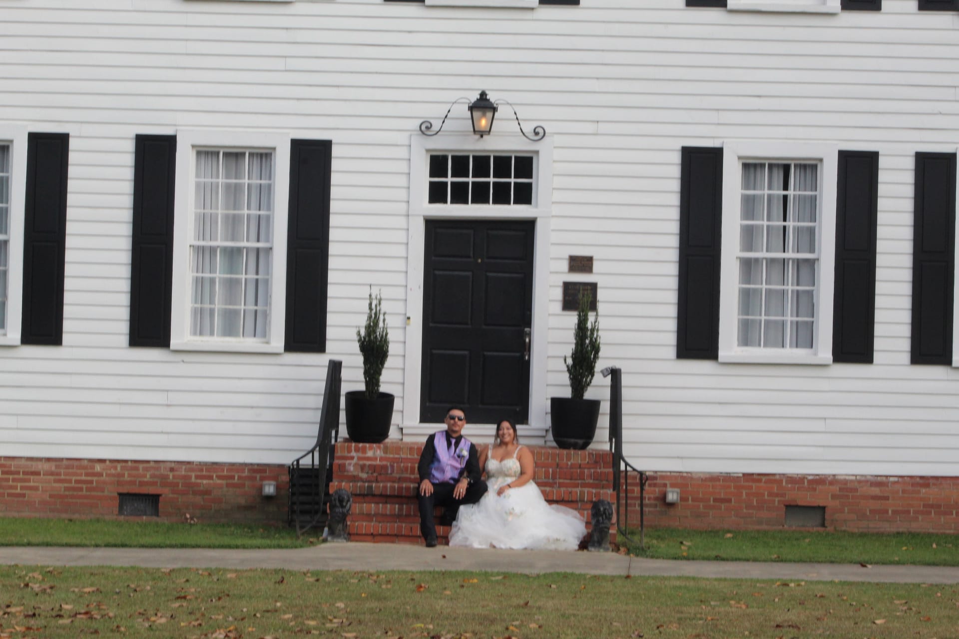 Bride in a white ballgown and groom in a black suit with lavender vest sit together on the brick front steps of Scarborough Houses 1821 white manor house with black shutters and a lantern-lit entrance door.