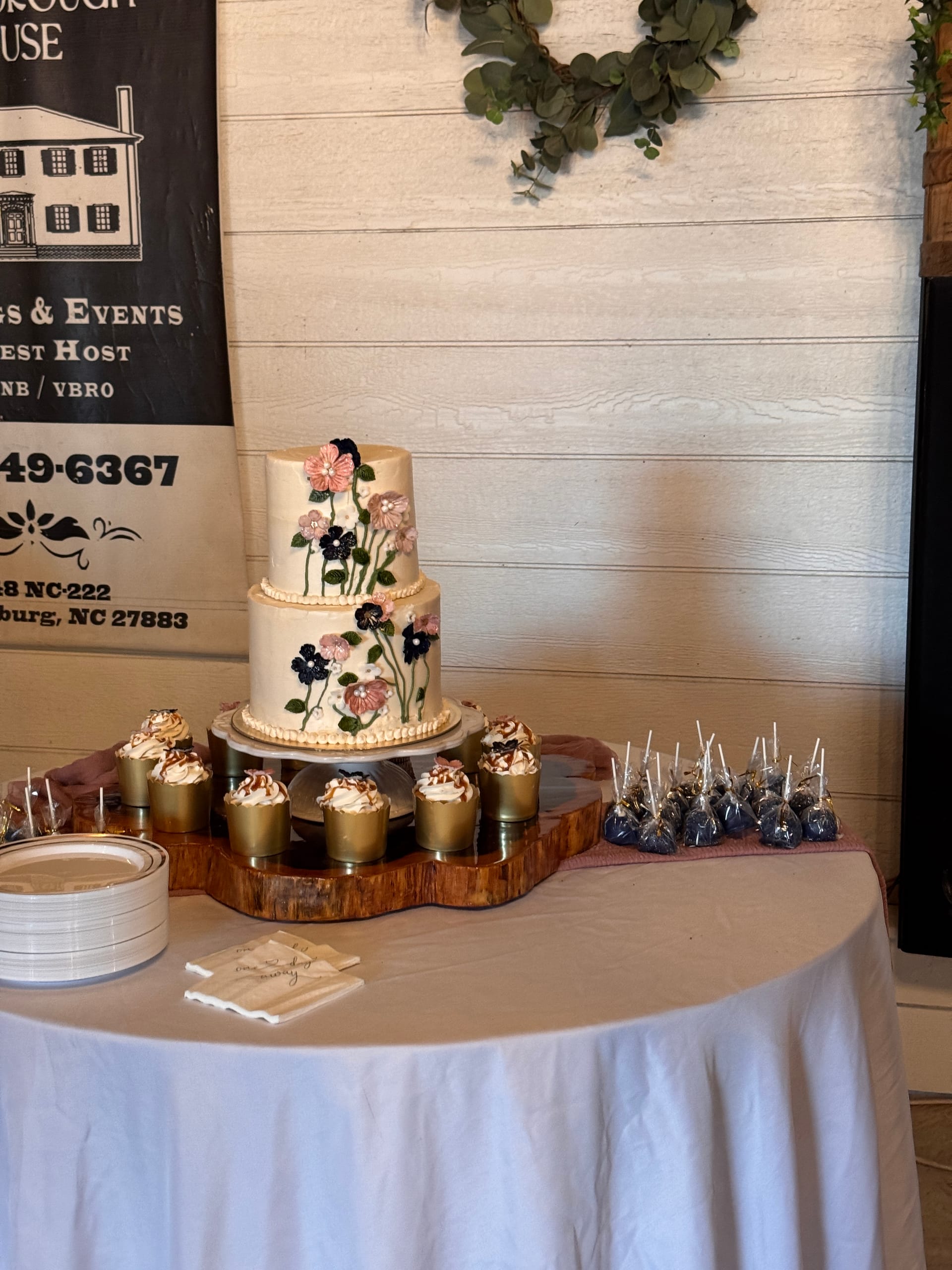 Elegant two-tier wedding cake adorned with hand-crafted pink and navy flowers displayed on a live-edge wood board surrounded by gold cupcakes and wrapped cake pops at Scarborough House venue in Stantonsburg NC.