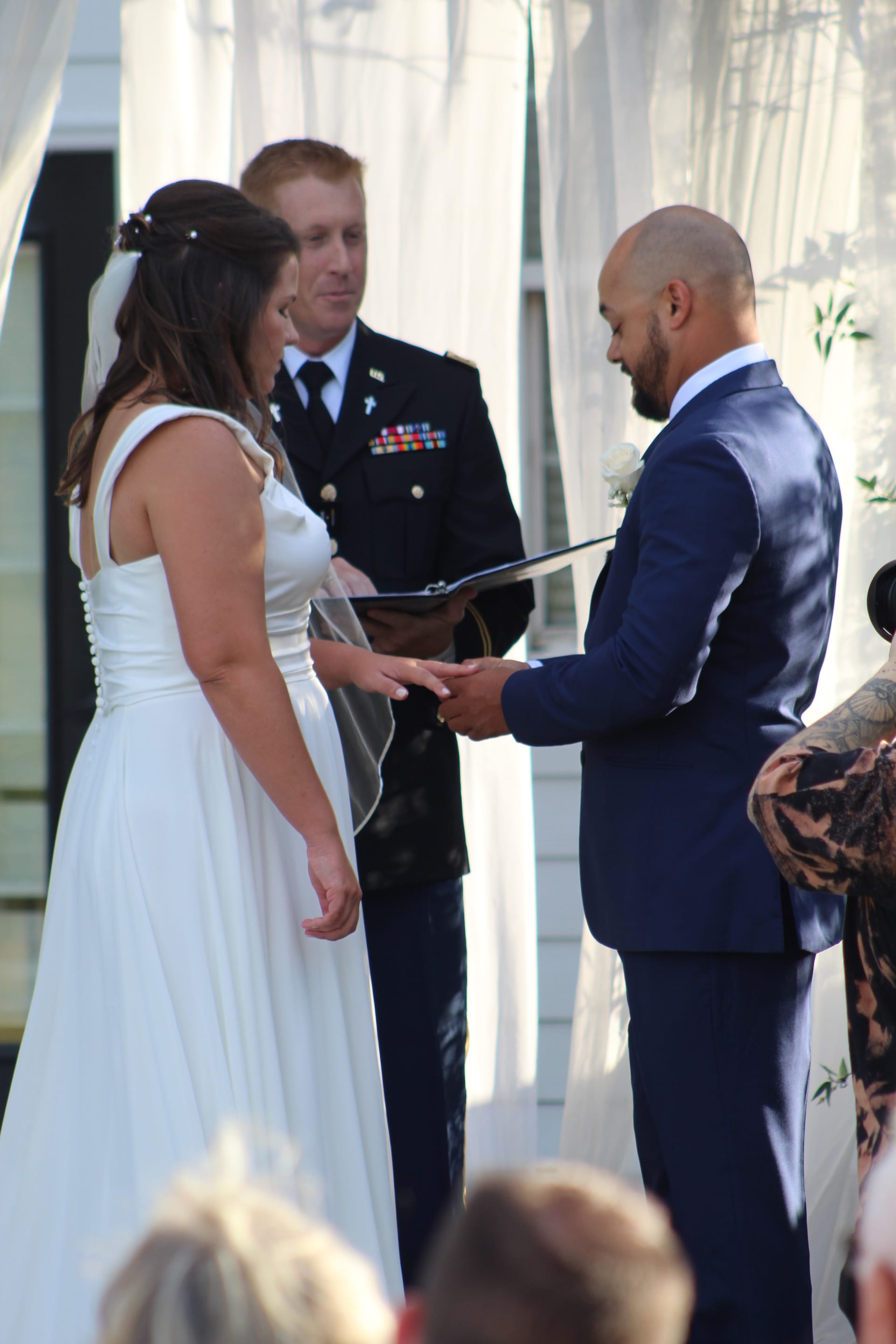 Groom in navy suit places a wedding ring on his brides finger during an outdoor ceremony officiated by a military officer at Scarborough House historic estate.