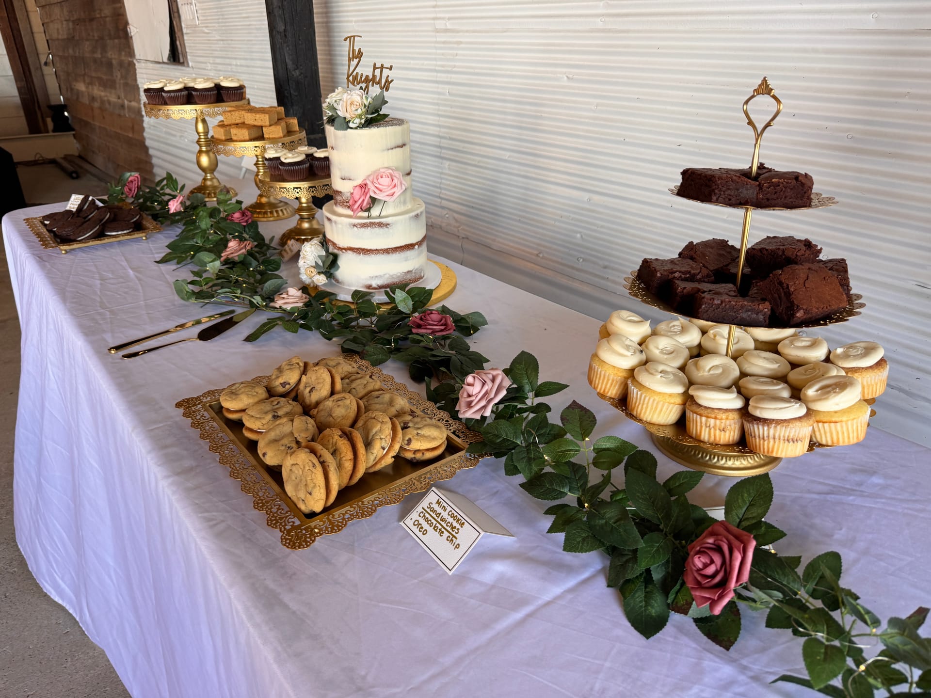 Elegant wedding dessert table featuring a two-tier semi-naked cake topped with pink roses and a gold The Knights topper, surrounded by vanilla cupcakes, brownies on a tiered stand, and chocolate chip cookie sandwiches on gold trays with a lush greenery and rose garland.