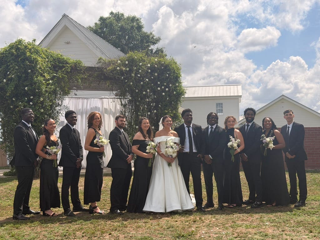 Bride and groom surrounded by their wedding party dressed in black standing on the ceremony lawn in front of a vine-covered white chapel at Scarborough House.