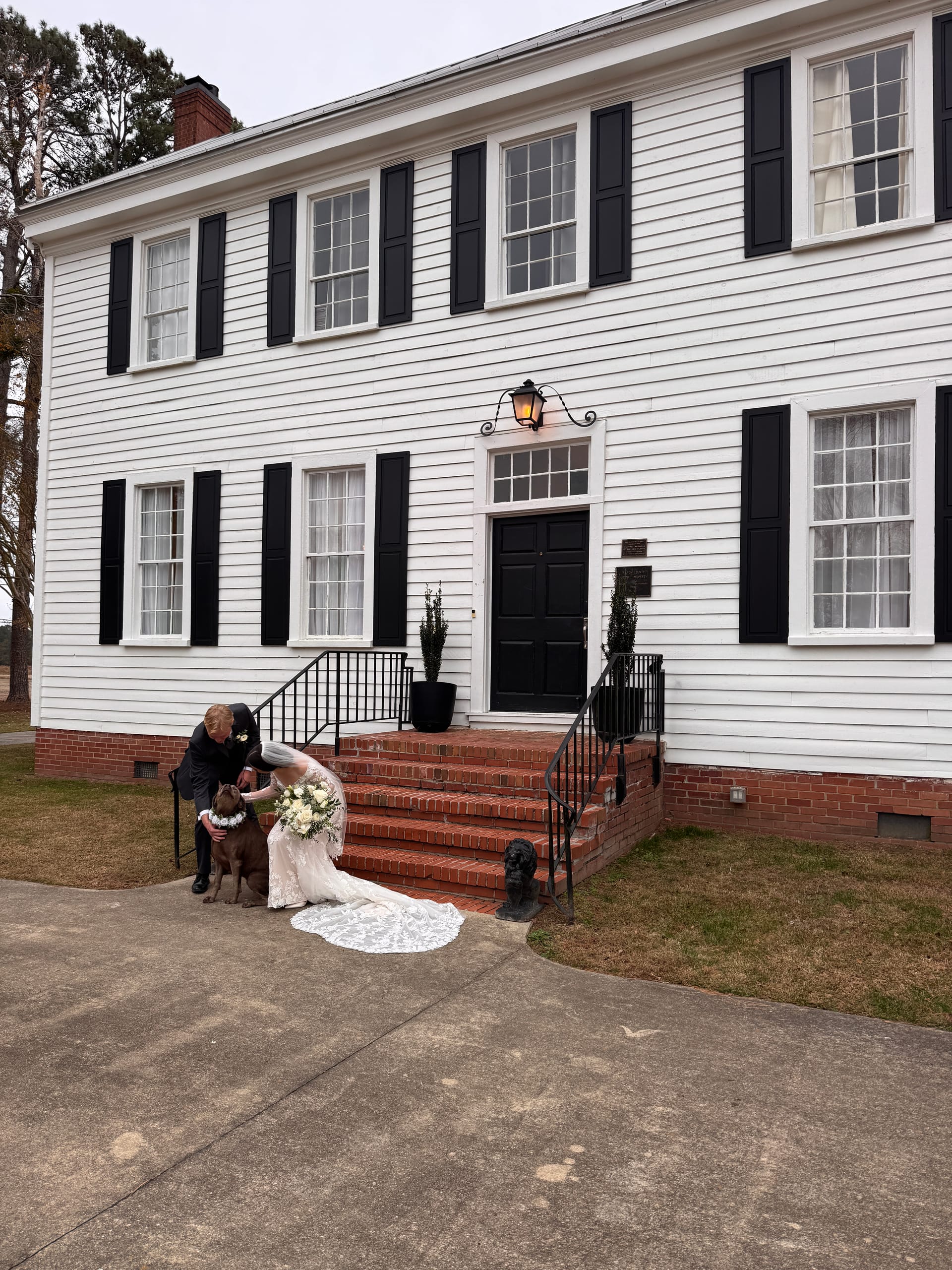 Bride in a lace gown kneeling to embrace her dog wearing a floral collar on the brick front steps of the 1821 Scarborough House manor while the groom looks on.