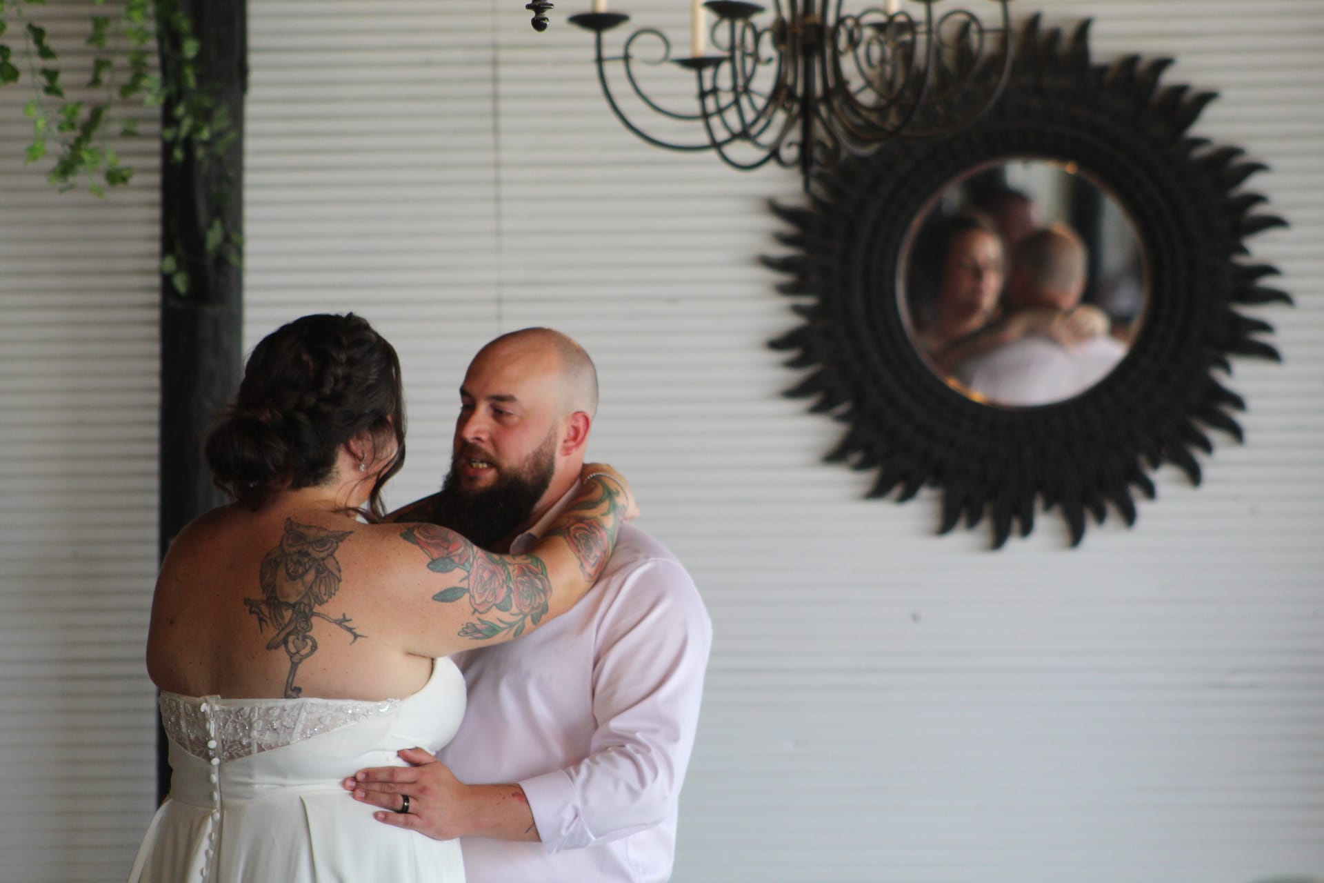 A tattooed bride and bearded groom share an intimate first dance inside Scarborough House grand hall, their embrace reflected in a decorative sunburst mirror beneath a wrought-iron chandelier.