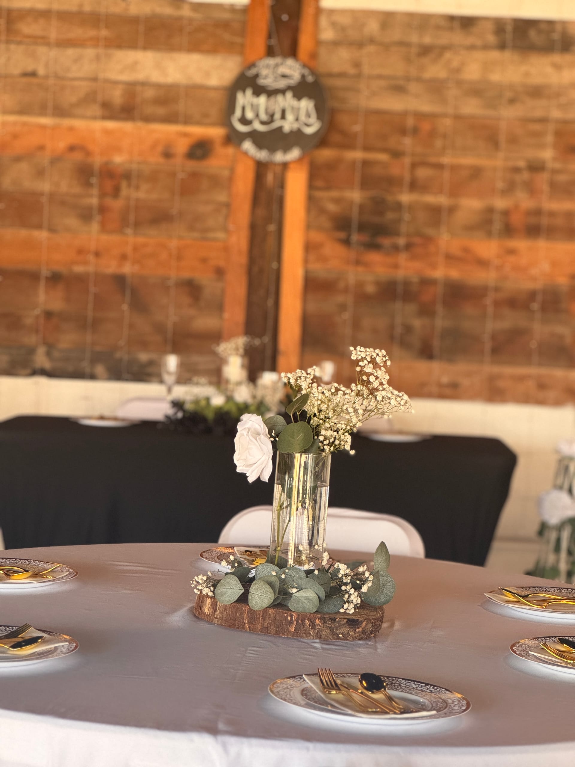 Rustic wedding reception table setting featuring white roses and baby's breath in glass vases on a wood slice centerpiece, surrounded by gold flatware on patterned plates, with a reclaimed wood accent wall in the background.