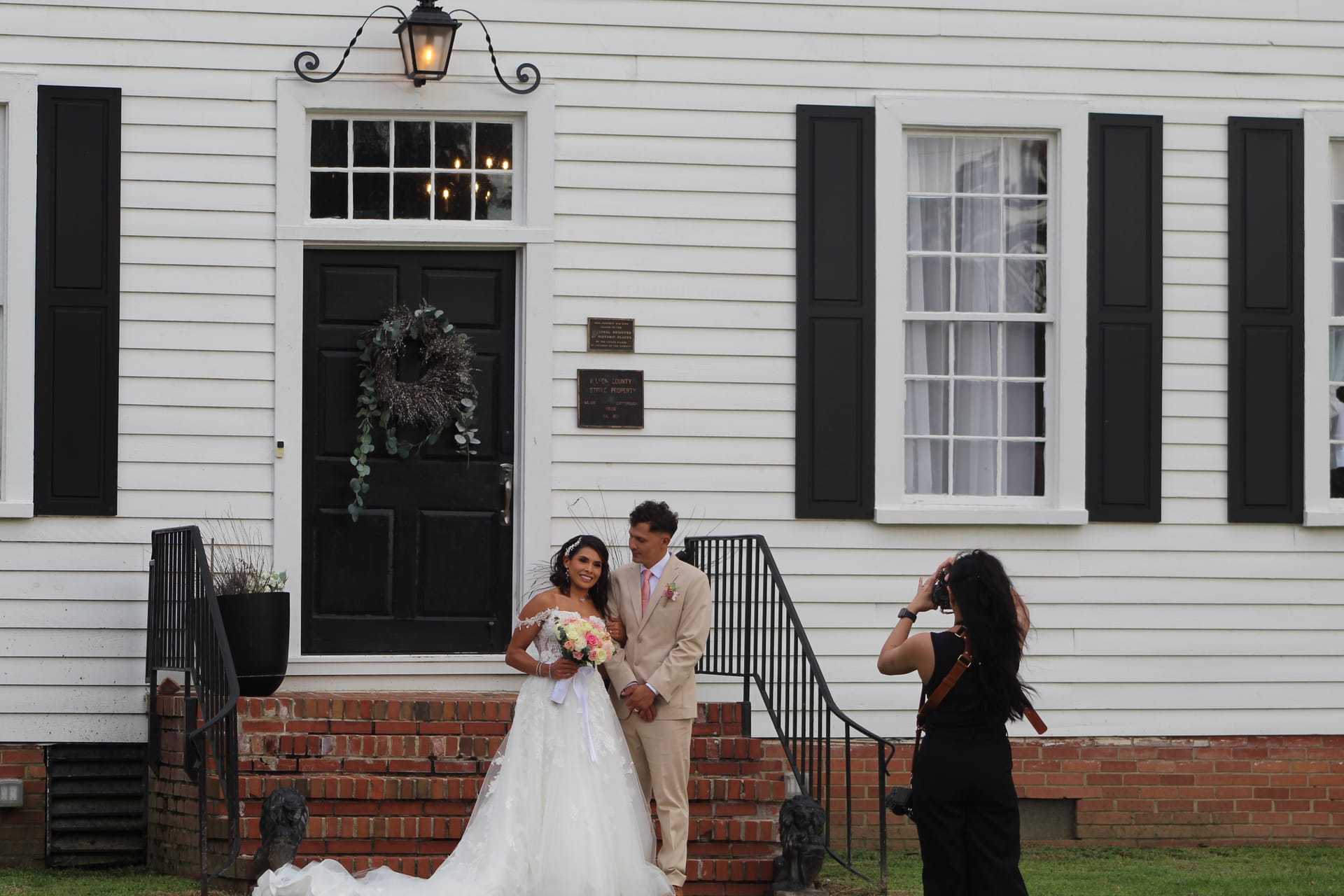 Bride in a white ball gown holding a pink bouquet and groom in a tan suit pose on the brick front steps of the 1821 Scarborough House manor while their photographer captures the moment.