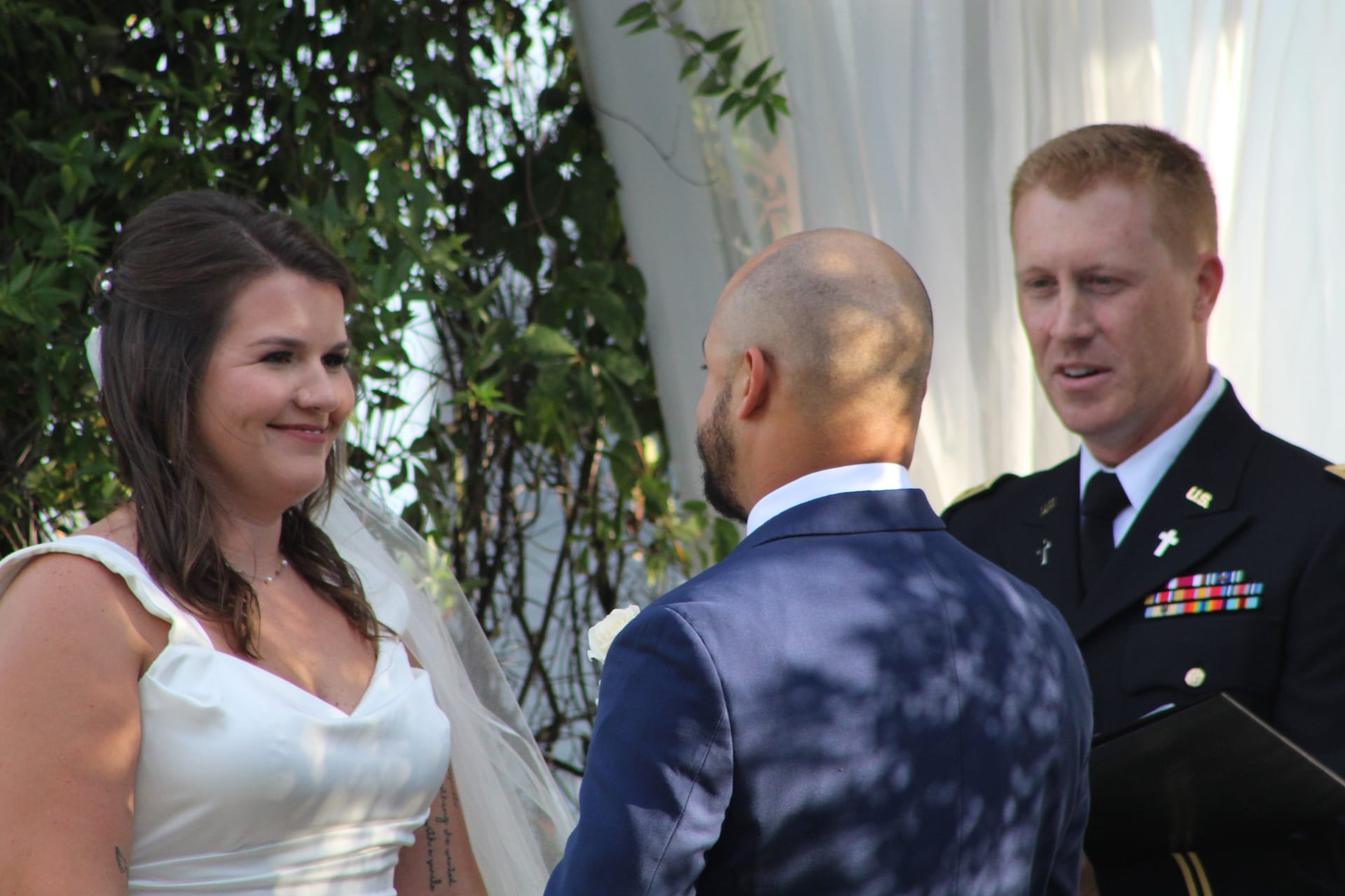 Bride in white gown and veil smiles warmly at her groom during outdoor wedding ceremony officiated by a military chaplain at Scarborough House estate.