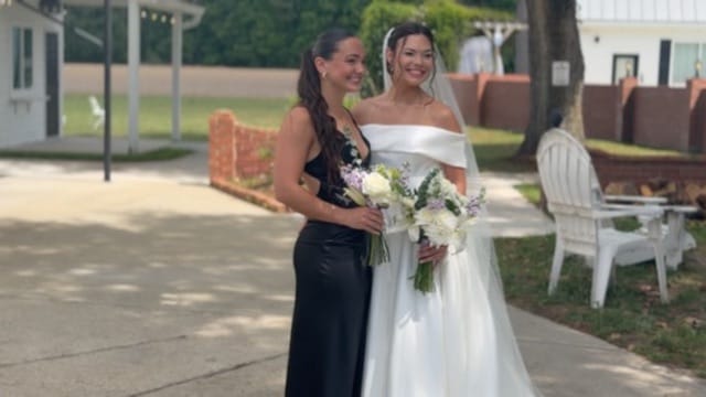 A smiling bride in an off-shoulder white gown holding a white and lavender bouquet poses with her bridesmaid in a black satin dress on the grounds of Scarborough House estate.