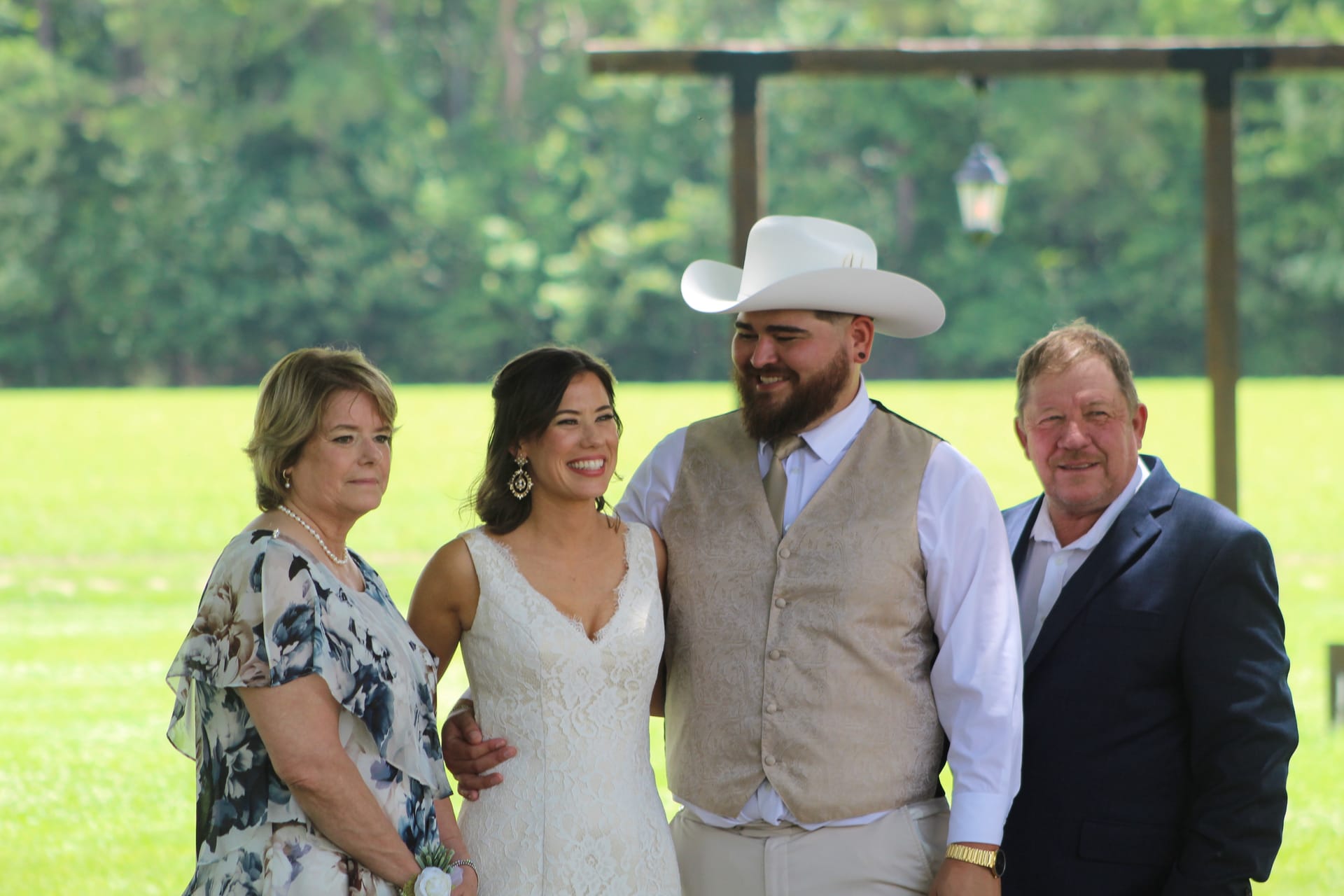 Bride in lace gown and groom in tan vest and white cowboy hat pose with two family members on the ceremony lawn at Scarborough House wedding venue.