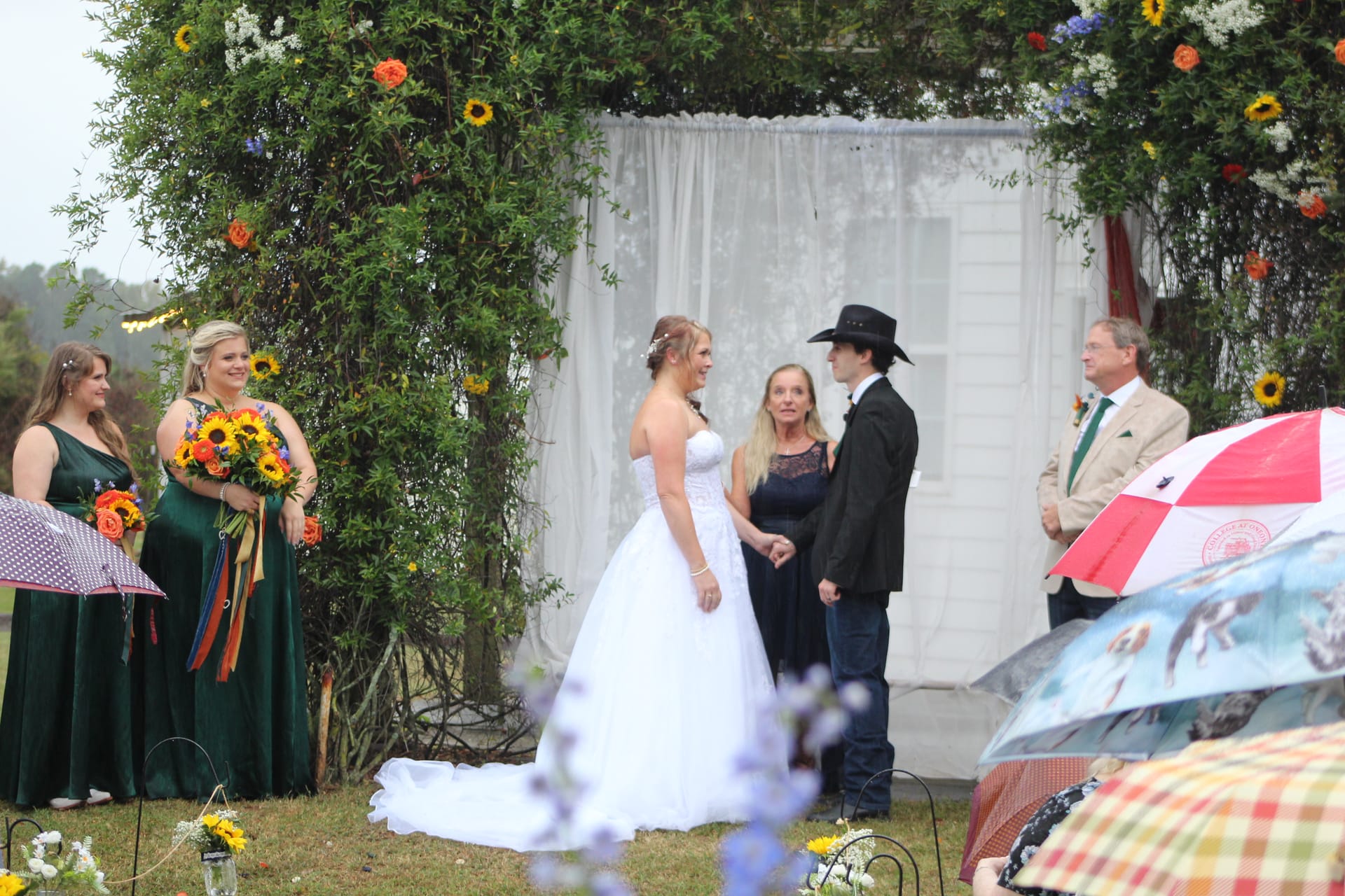 Bride and groom exchange vows beneath a lush floral arch decorated with sunflowers and wildflowers on the ceremony lawn at Scarborough House while bridesmaids in emerald green look on during a light rain.