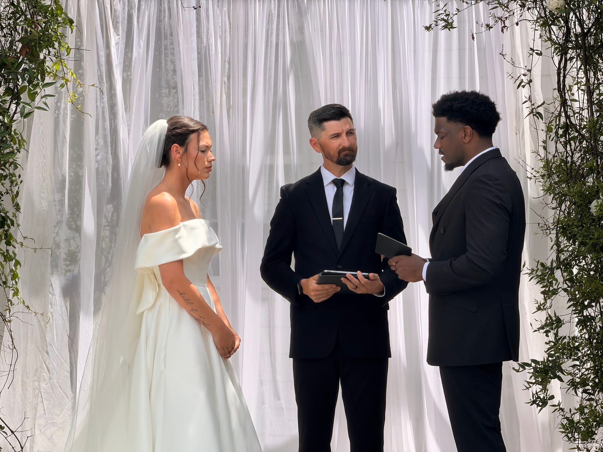 Bride in an off-shoulder white gown and groom in a black suit exchange vows with their officiant beneath a white draped altar adorned with greenery at Scarborough House.