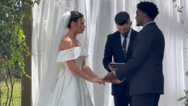Bride in an off-shoulder white gown and groom in a black suit exchange vows before an officiant against a flowing white drapery backdrop at a garden ceremony.