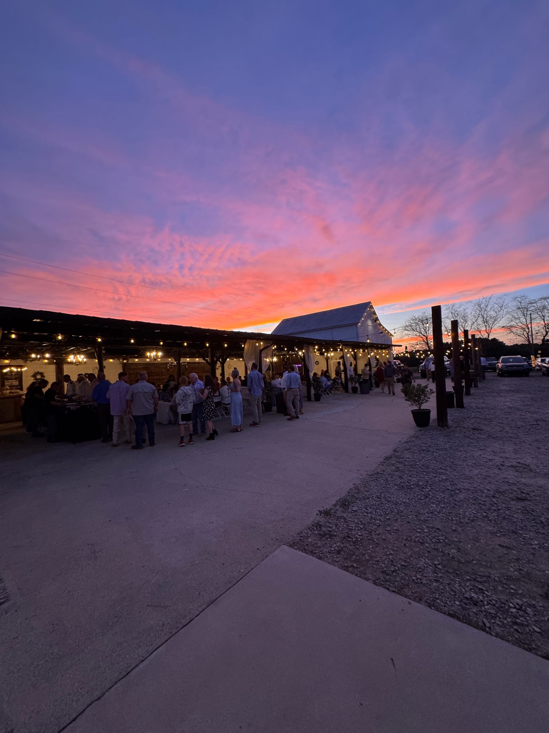 Guests mingle under string lights at an outdoor wedding reception pavilion bathed in a breathtaking pink and purple sunset sky.