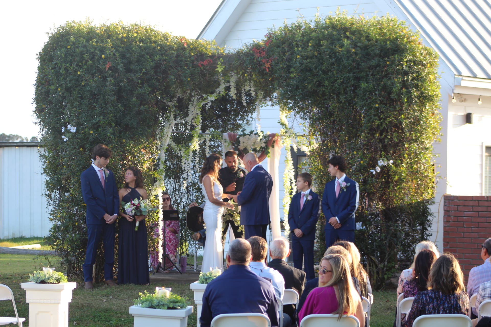 A bride and groom exchange vows beneath a lush green hedge arch adorned with white florals on the ceremony lawn at Scarborough House, surrounded by their wedding party and seated guests.