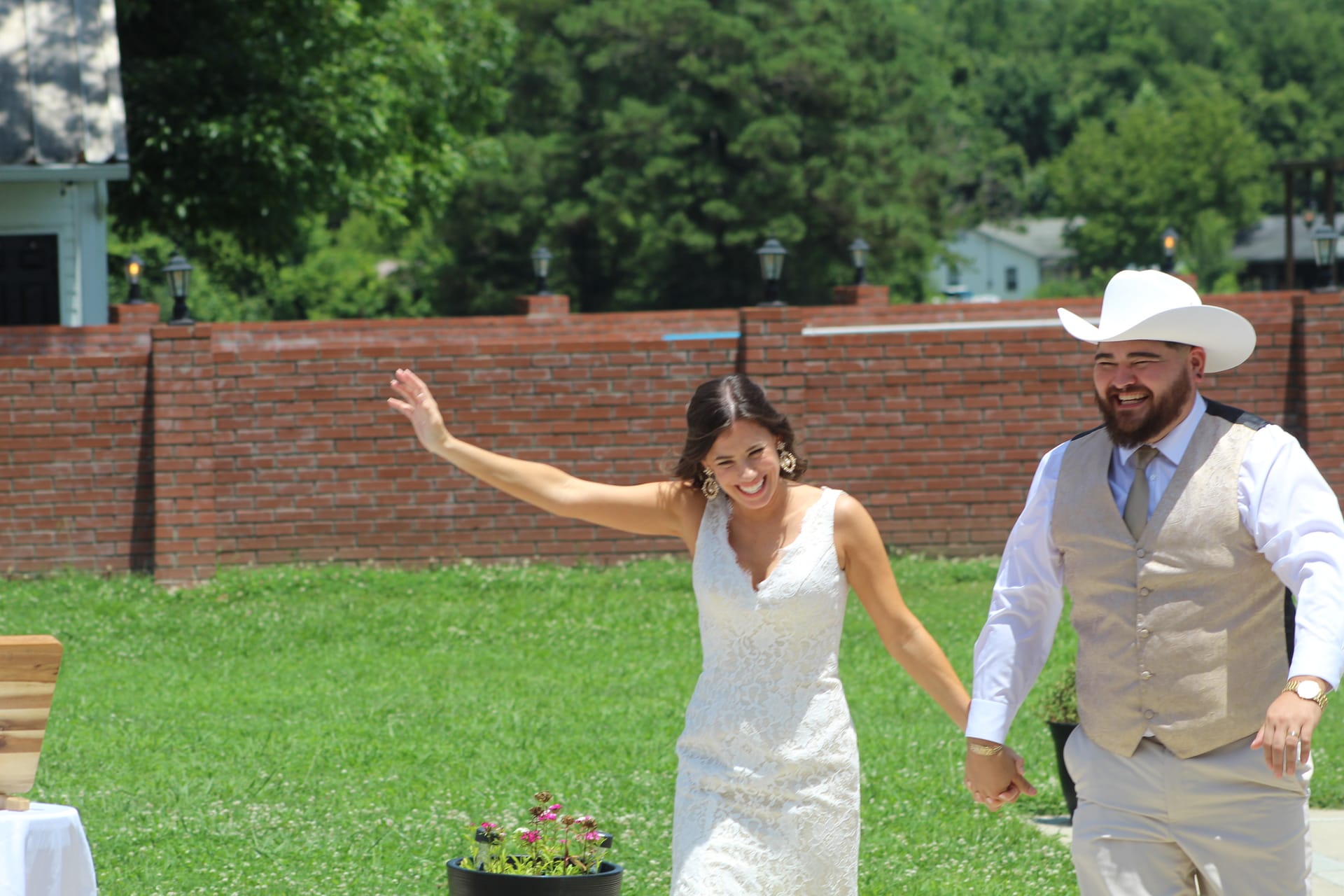 Smiling bride in white lace gown waves while holding hands with bearded groom in white cowboy hat and tan vest as they make their grand entrance on the sunlit ceremony lawn at Scarborough House.