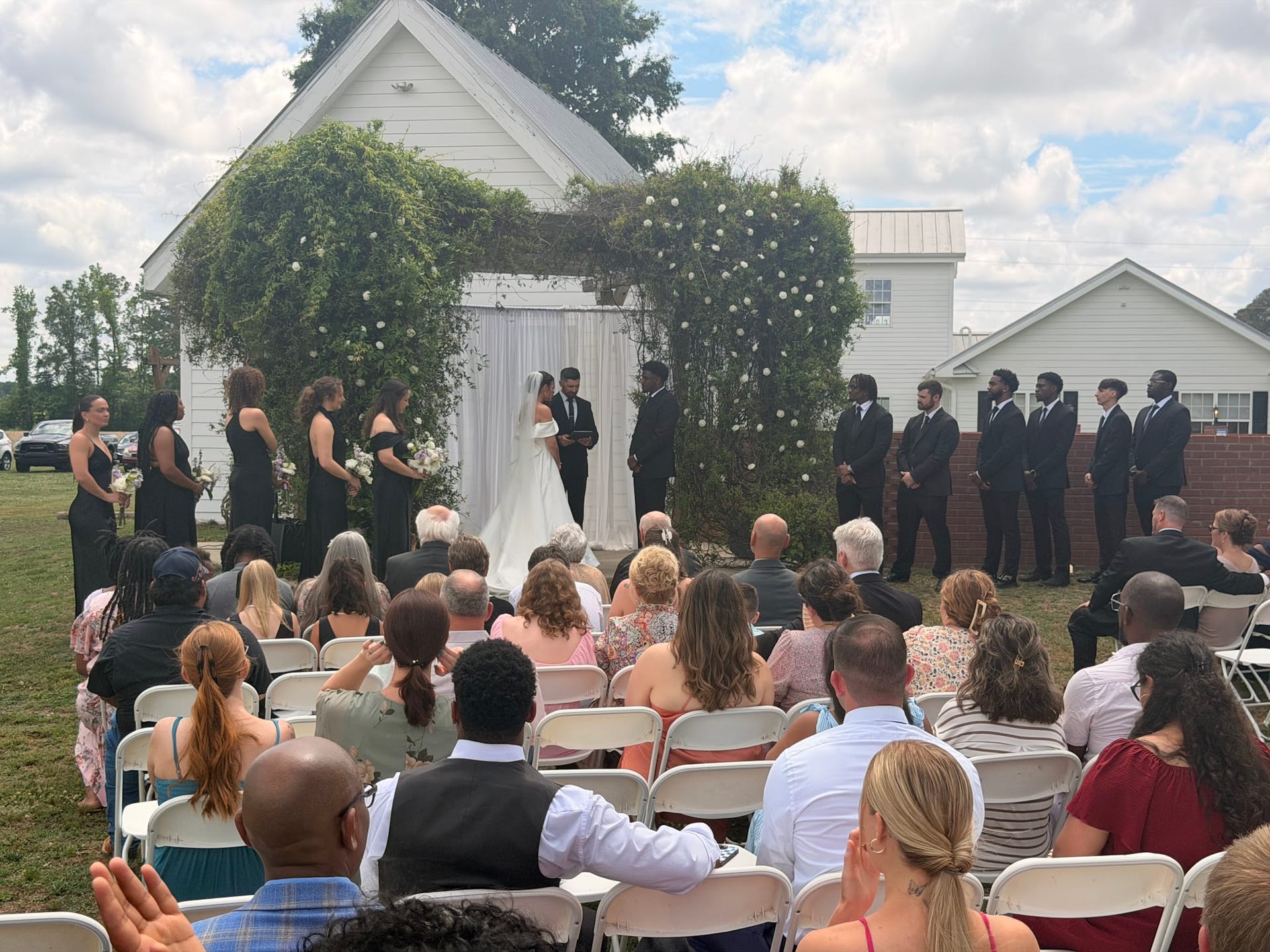 Outdoor wedding ceremony at Scarborough House with bride and groom exchanging vows beneath a lush floral arch flanked by bridesmaids in black and groomsmen in suits while guests watch from white folding chairs on the estate lawn.