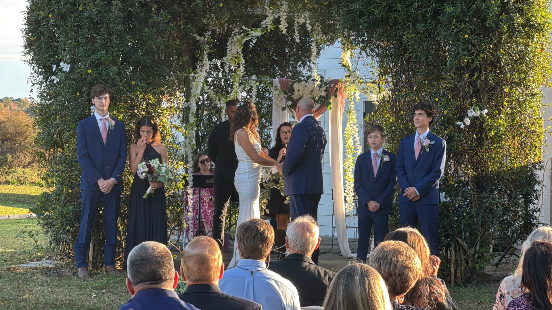 Bride and groom exchange vows beneath a lush floral arch on the ceremony lawn at Scarborough House historic estate, surrounded by their wedding party and seated guests in warm afternoon light.