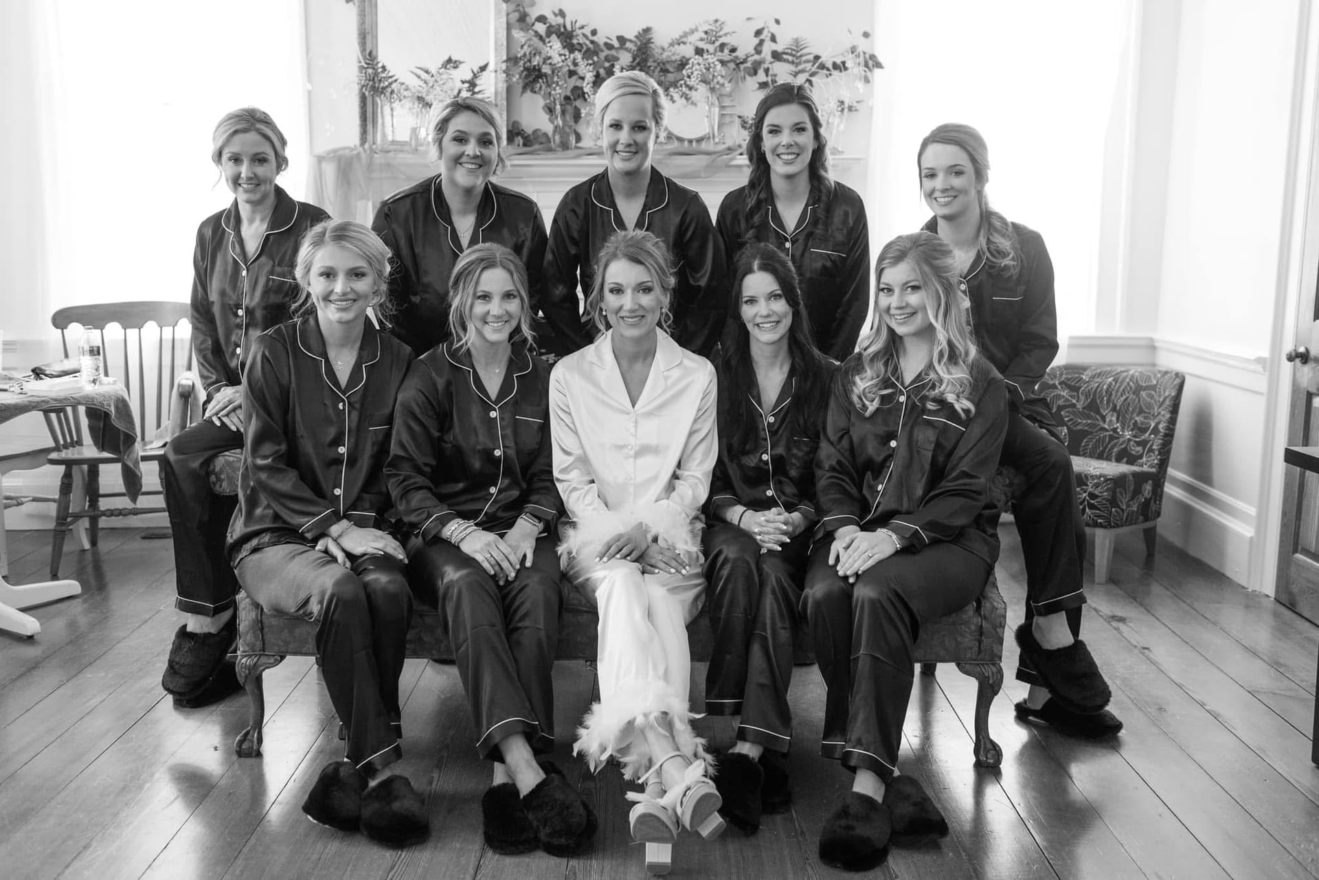 Bride in white satin pajamas seated at center surrounded by nine bridesmaids in matching dark satin sets posing together in a warmly lit historic room at Scarborough House.