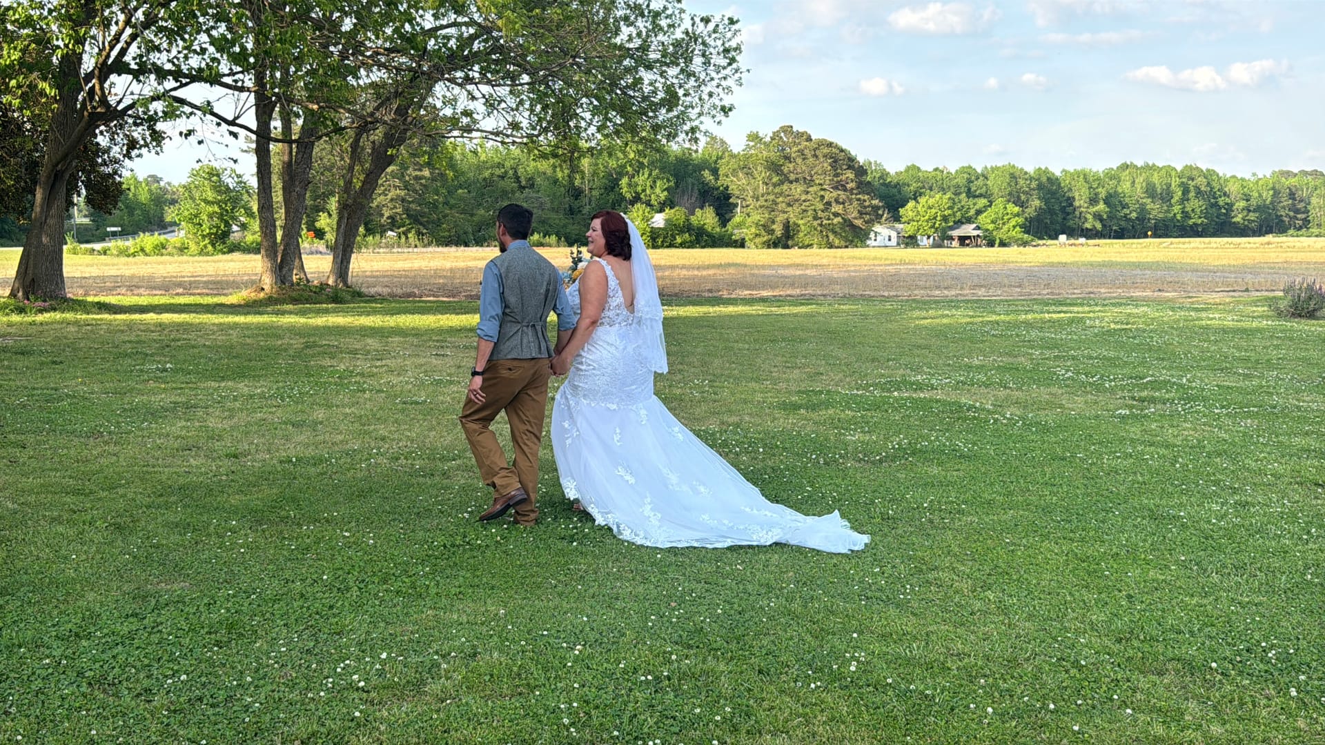 Bride in a lace gown with cathedral train glances back while walking hand in hand with groom across a sunlit green lawn with open farmland and trees in the background.