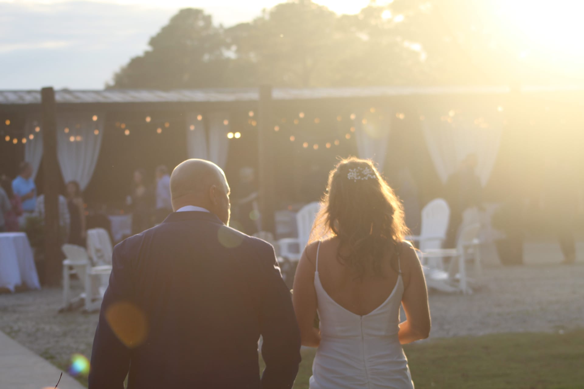 Bride and groom walk hand in hand toward a string-light adorned pavilion bathed in warm golden sunset light at Scarborough House.