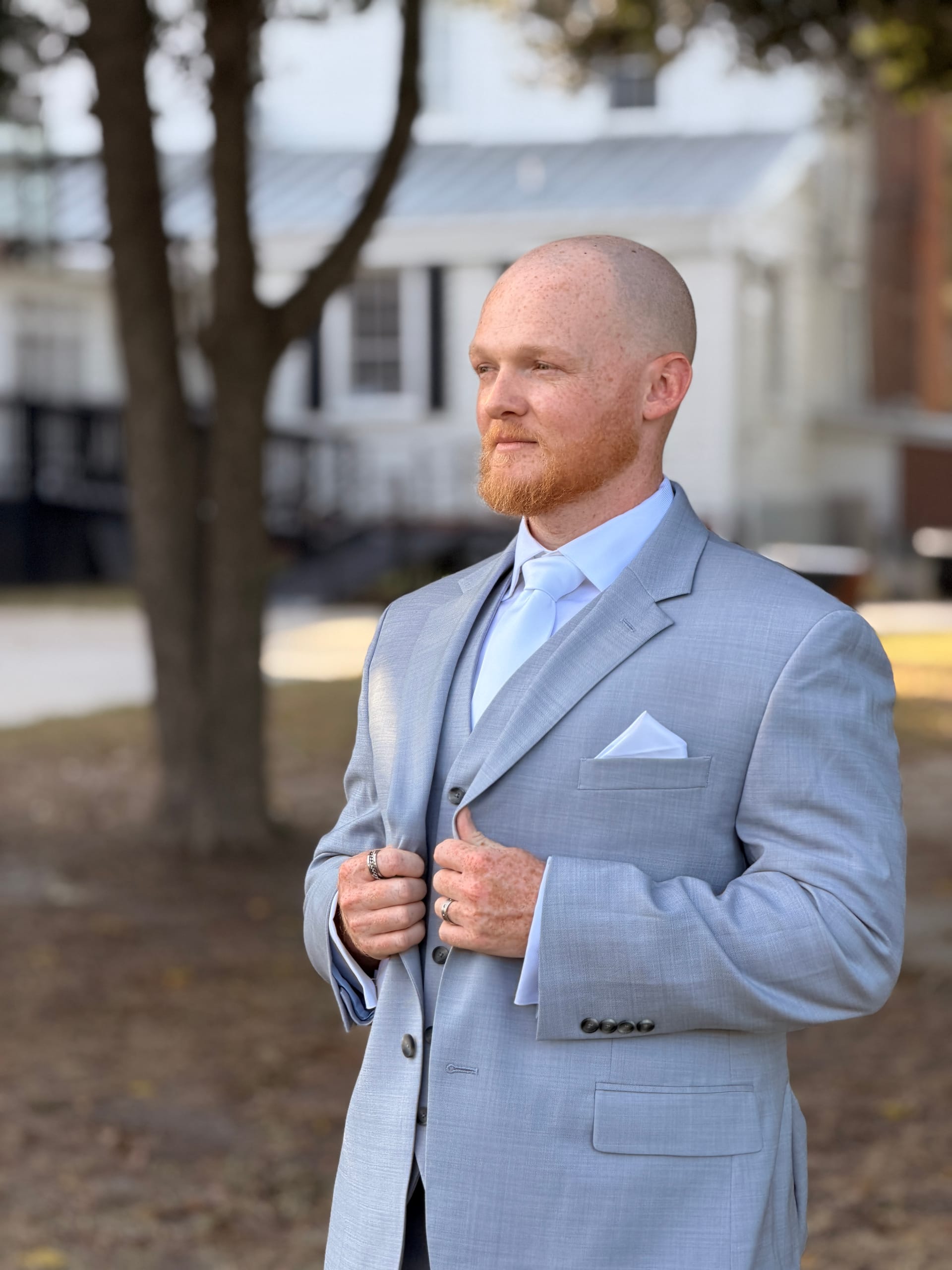 Bald groom with a red beard wearing a light grey three-piece suit and white tie stands confidently outdoors among oak trees at Scarborough House historic wedding venue.